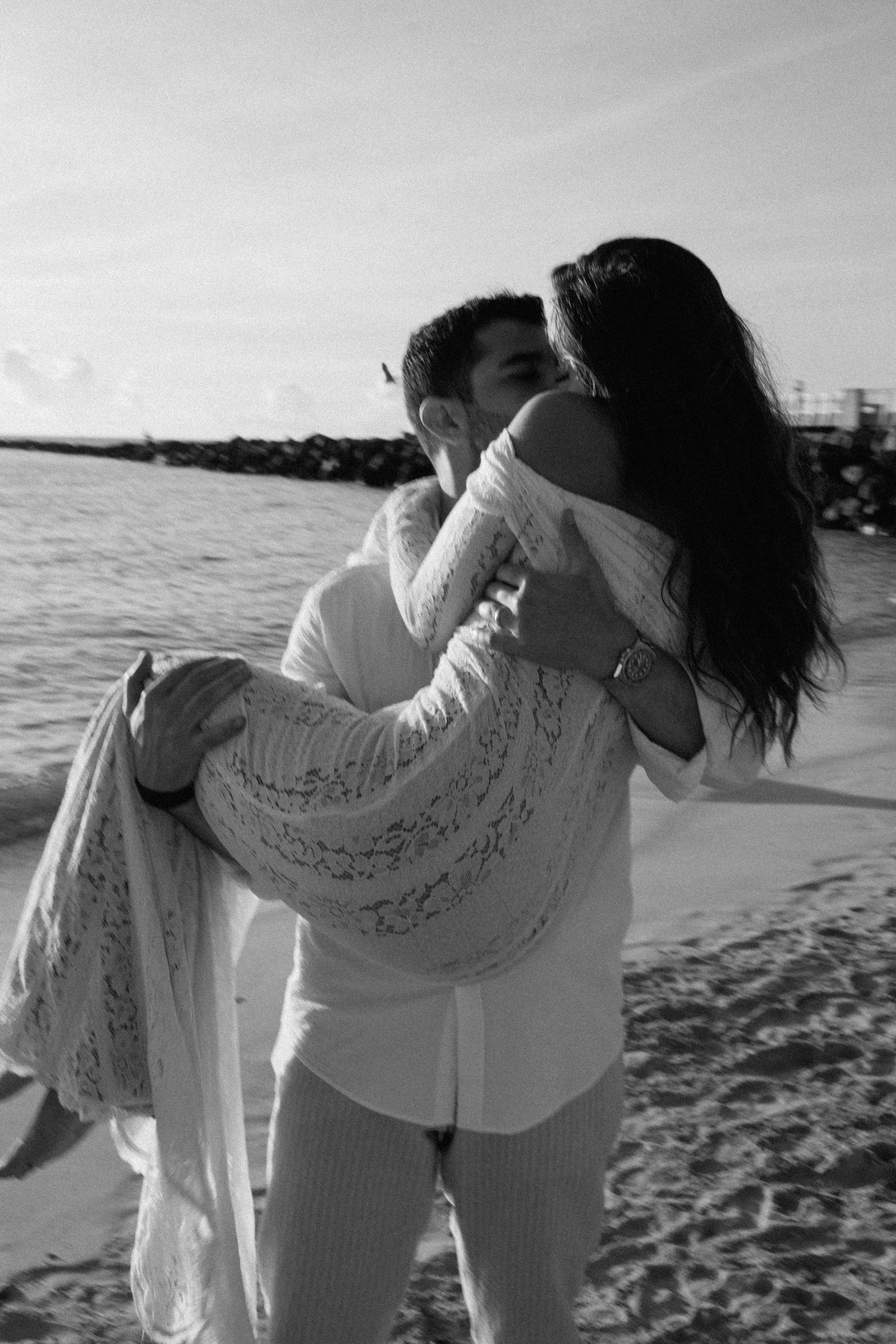A black and white beach photo of a couple sharing a tender moment as waves crash nearby.