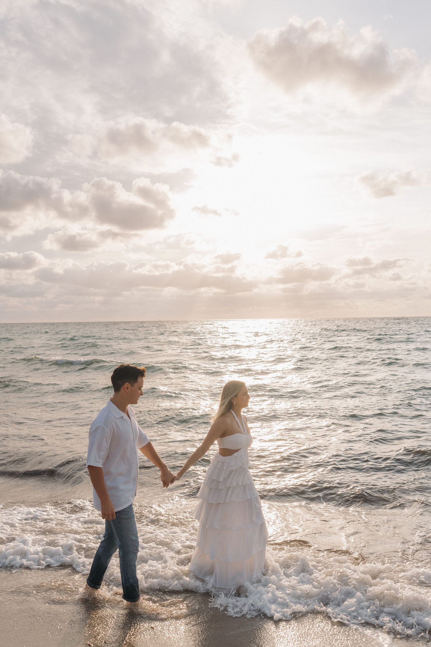 Couple walking together through ocean waves during sunset with soft clouds overhead.