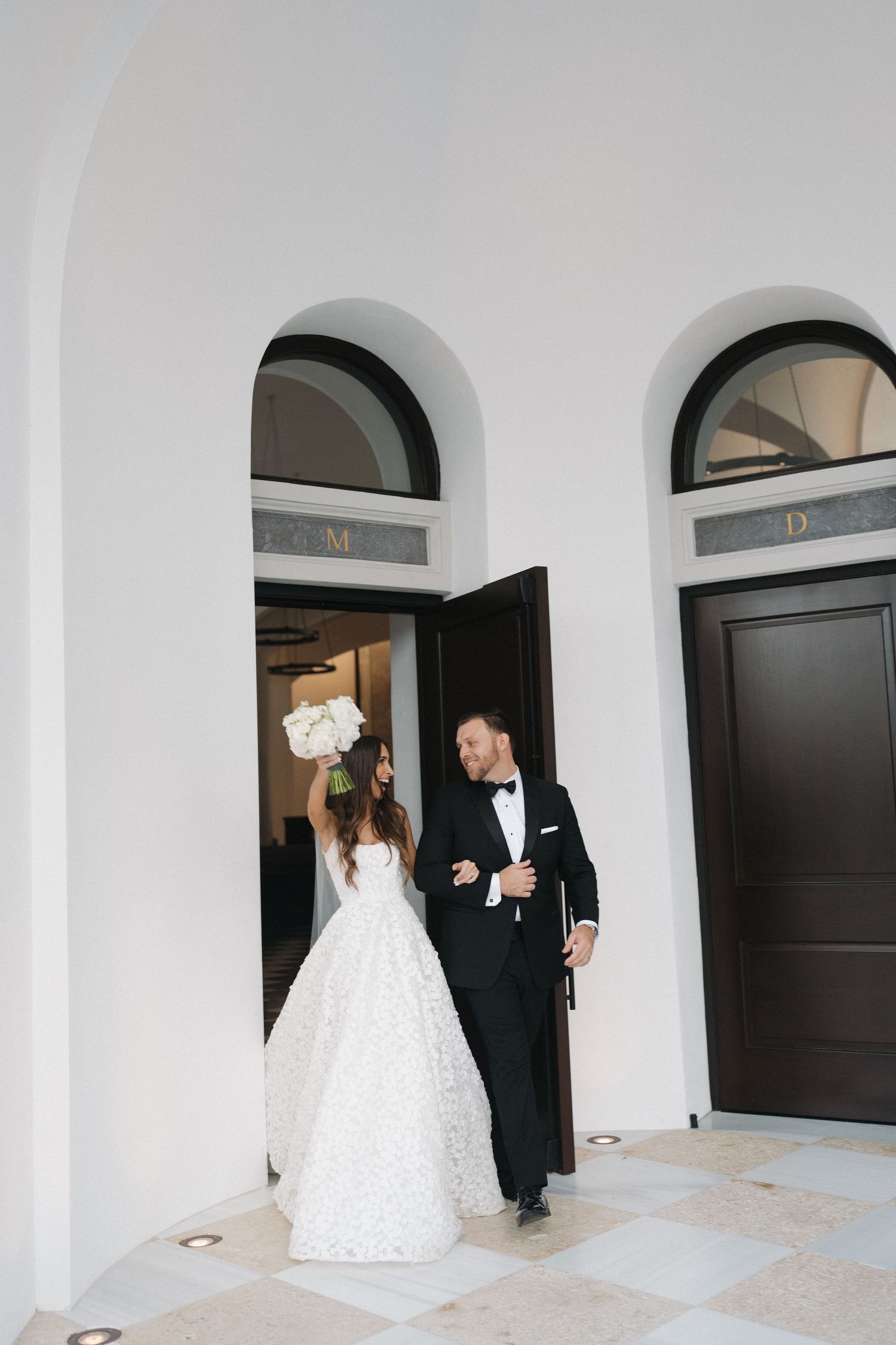 A couple walks through an elegant white hallway with arched doorways.