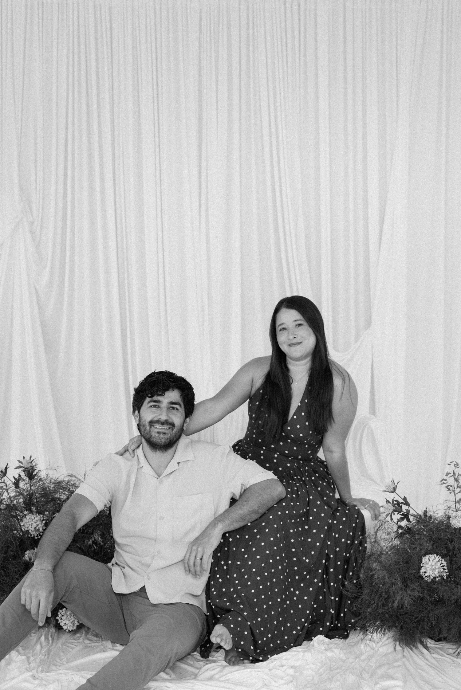Black and white photo of a smiling couple posing against white draped curtains with floral arrangements.