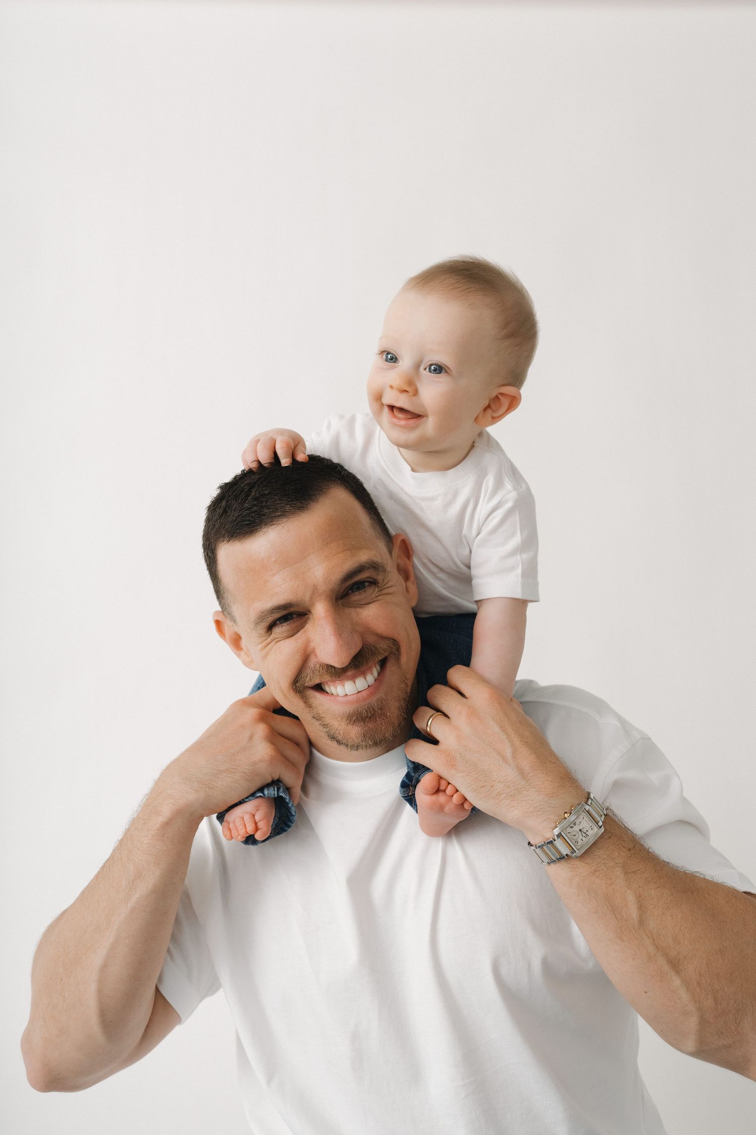 Baby riding on parent's shoulders against white background.