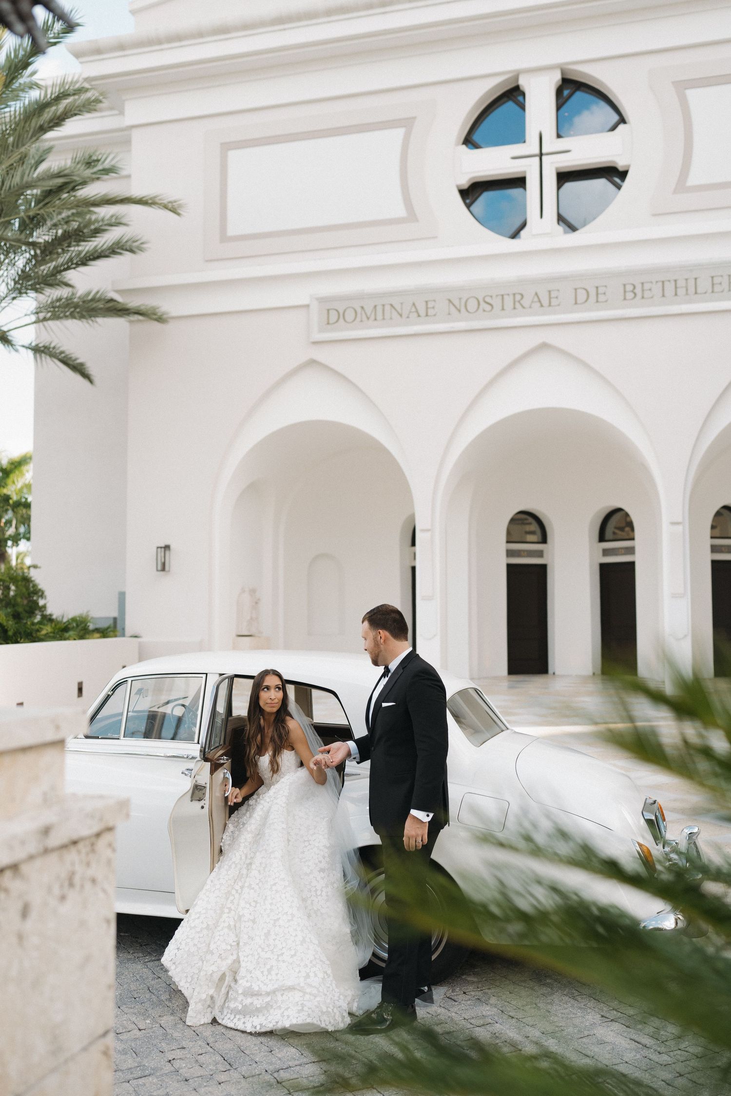 A couple poses in front of a white church with palm trees and architectural details.