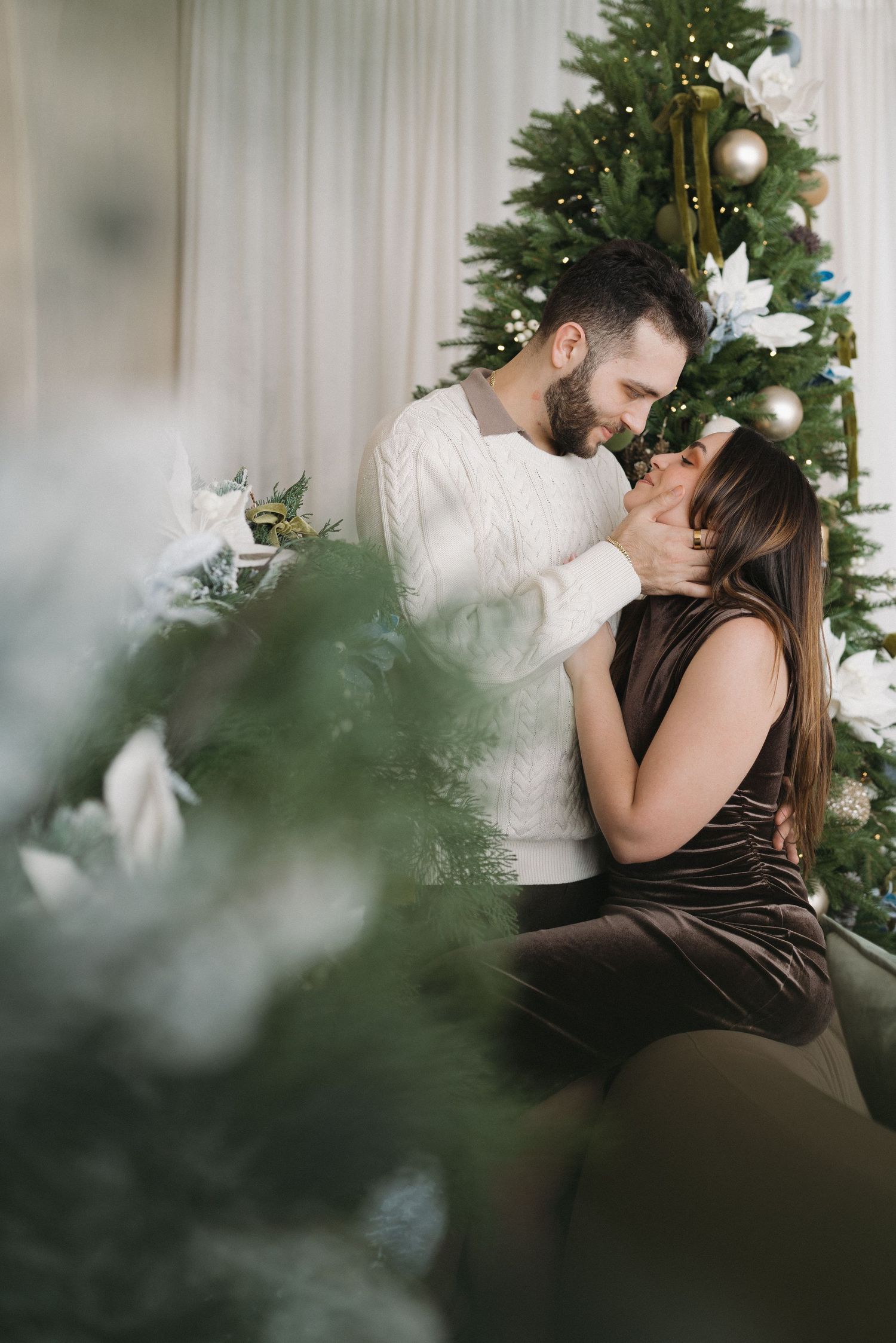 Romantic couple sharing a tender moment near a decorated Christmas tree, man in white sweater, woman in brown velvet dress.