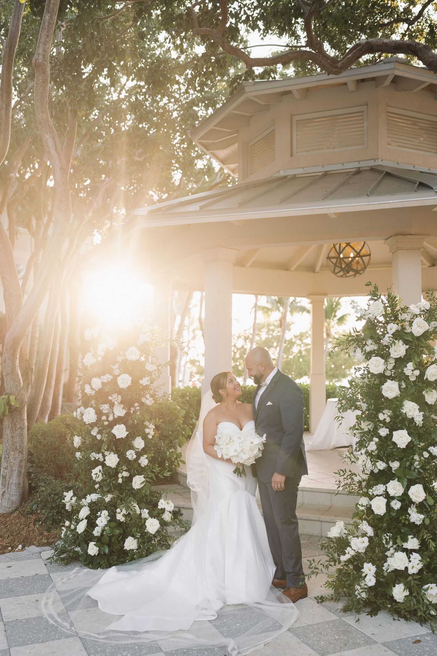 A couple in wedding attire poses in front of white roses and a garden gazebo during golden hour.