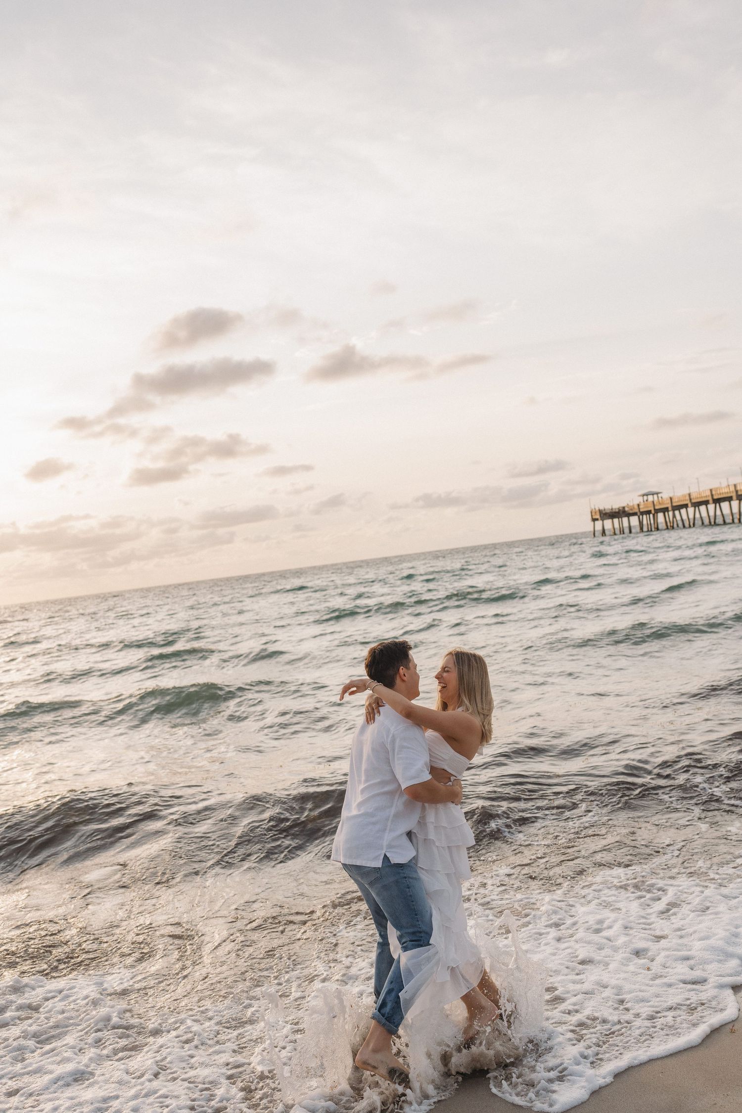 Couple sharing an intimate moment at the beach with a pier in the background during golden hour.