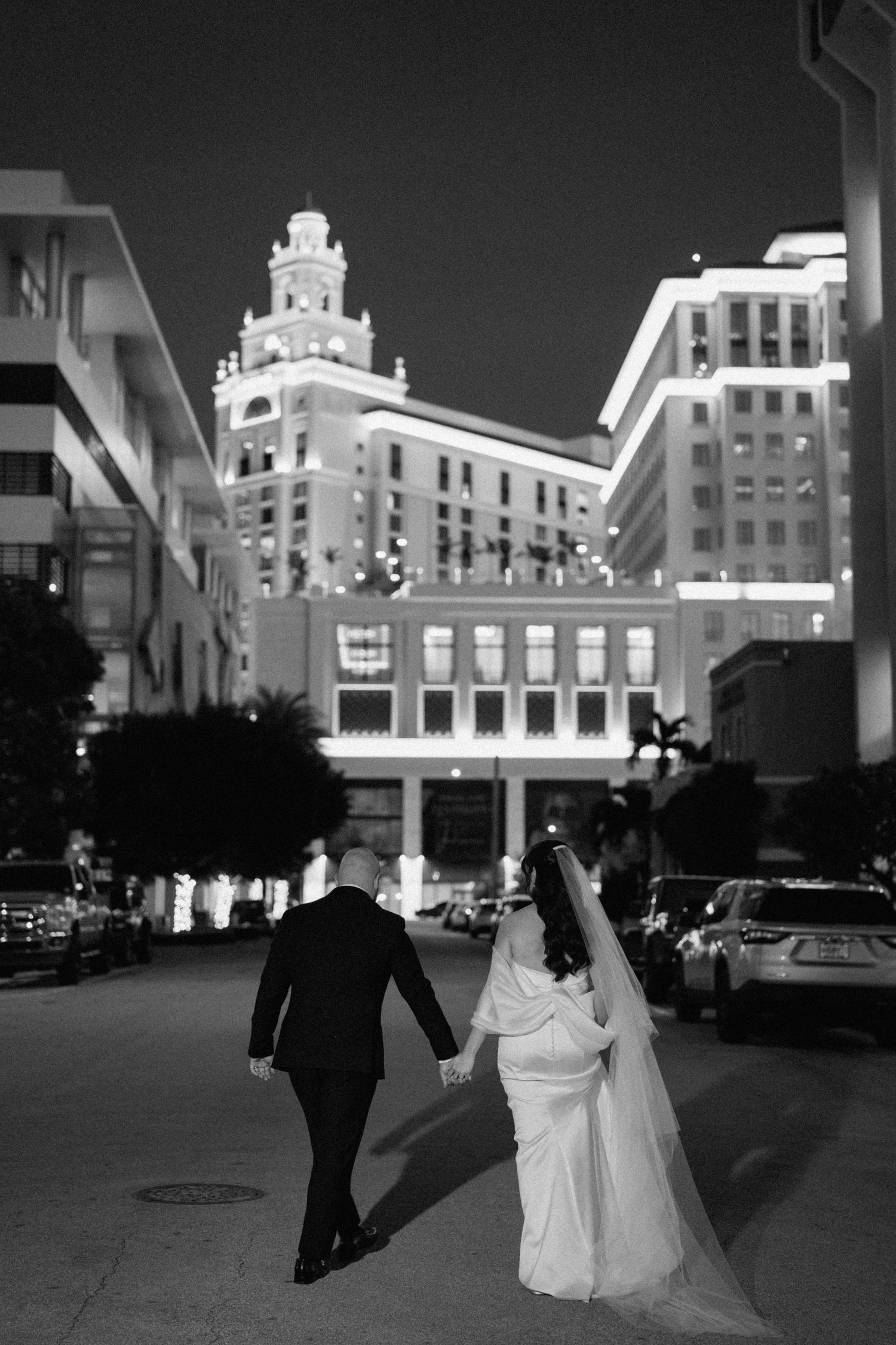 A couple walks hand in hand at night with illuminated art deco buildings in the background.