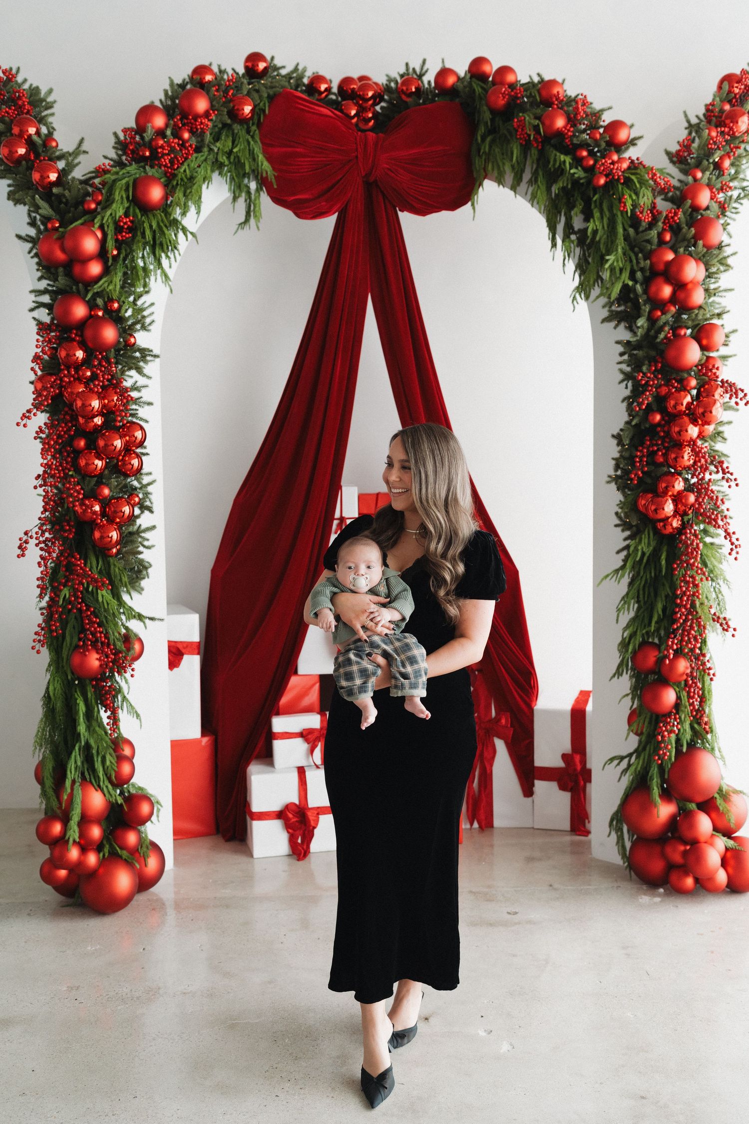 Woman in black dress holding baby under festive Christmas arch with red ornaments, velvet bow, and gift boxes.