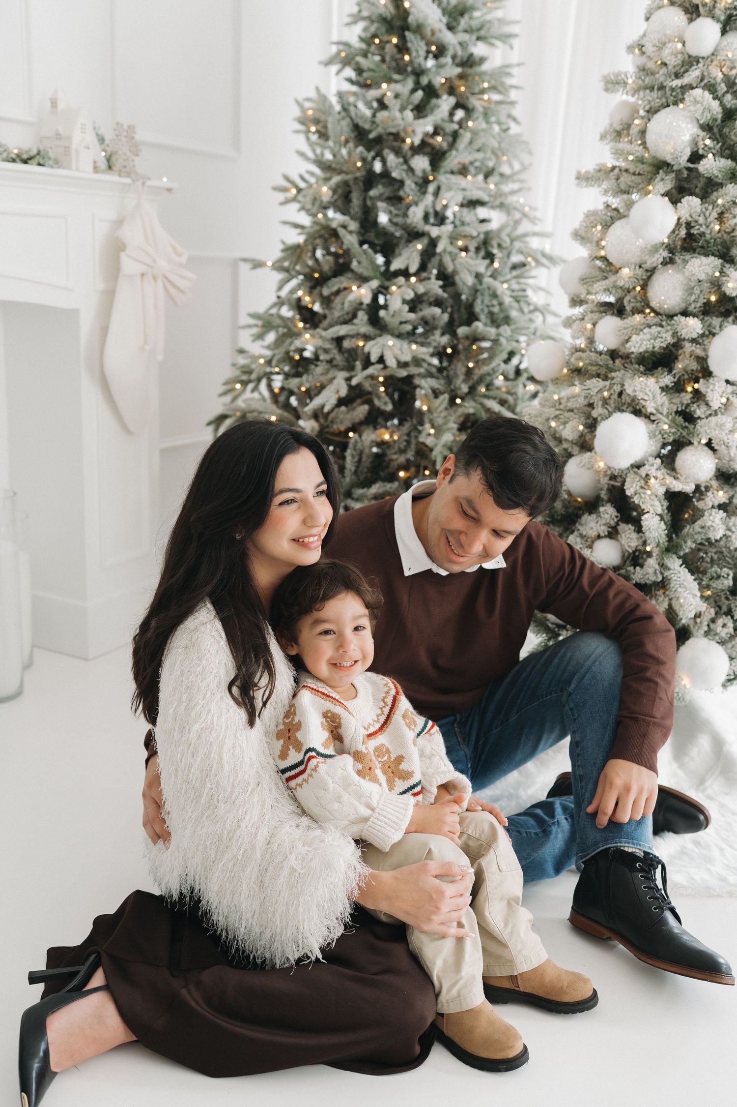 Happy family sitting together by a decorated Christmas tree in a white room, mom hugging child, dad smiling nearby.
