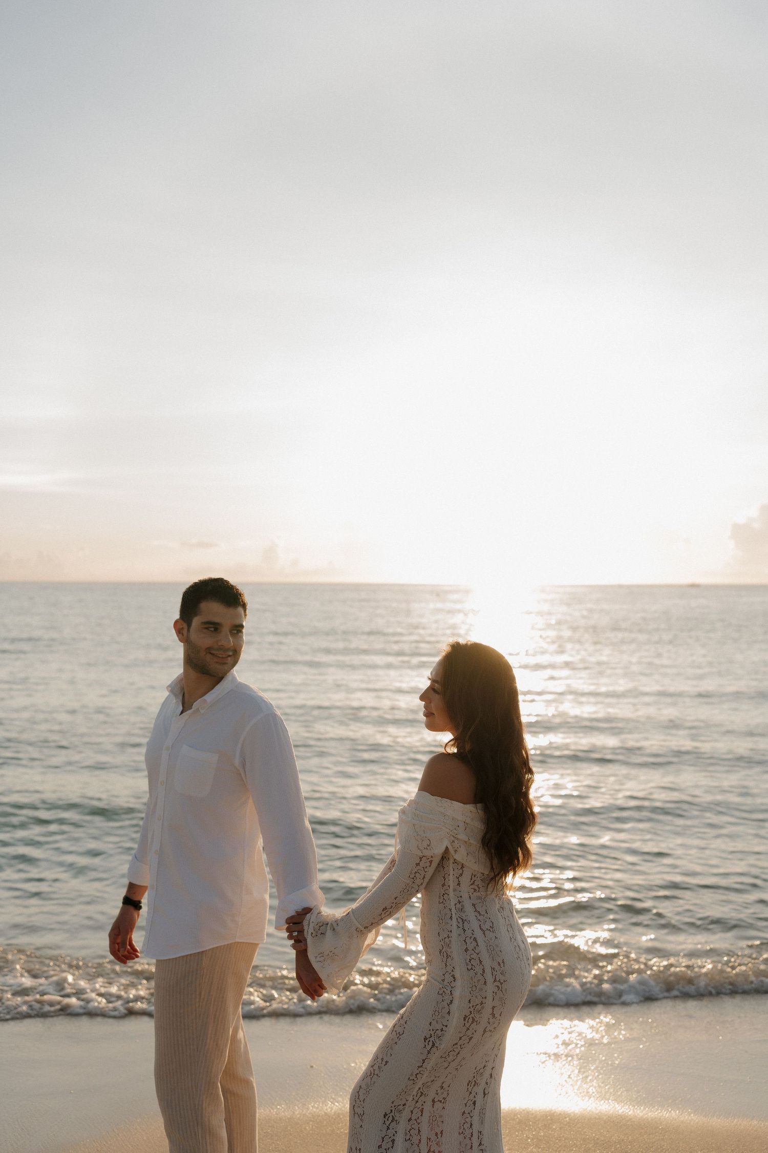 A couple in white outfits holds hands while walking along a serene beach at sunset.