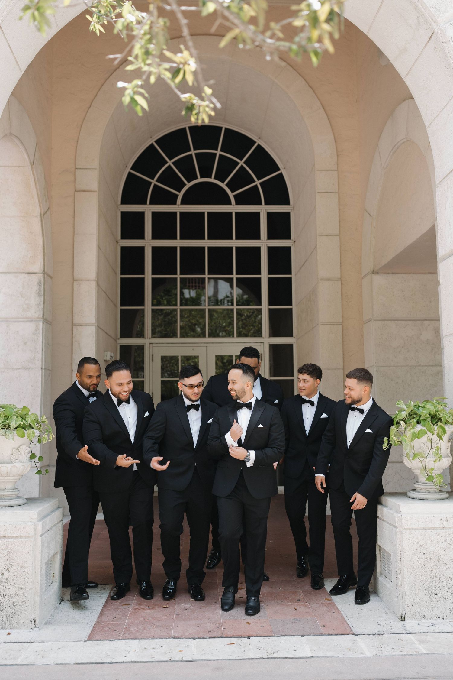 Groomsmen in black tuxedos gather under arched entrance for wedding photo.