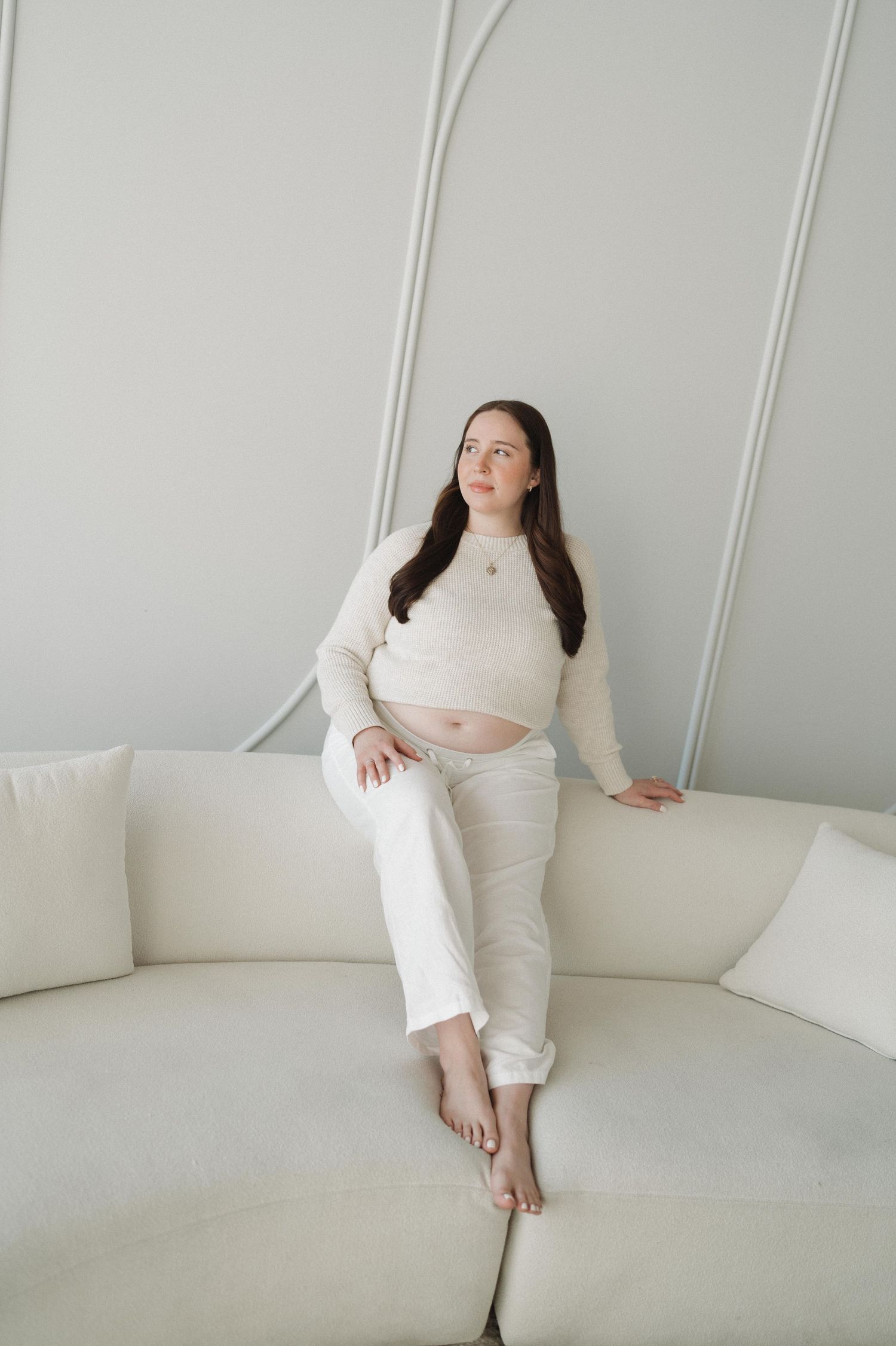 Relaxed maternity pose on white couch featuring cream-colored casual outfit.