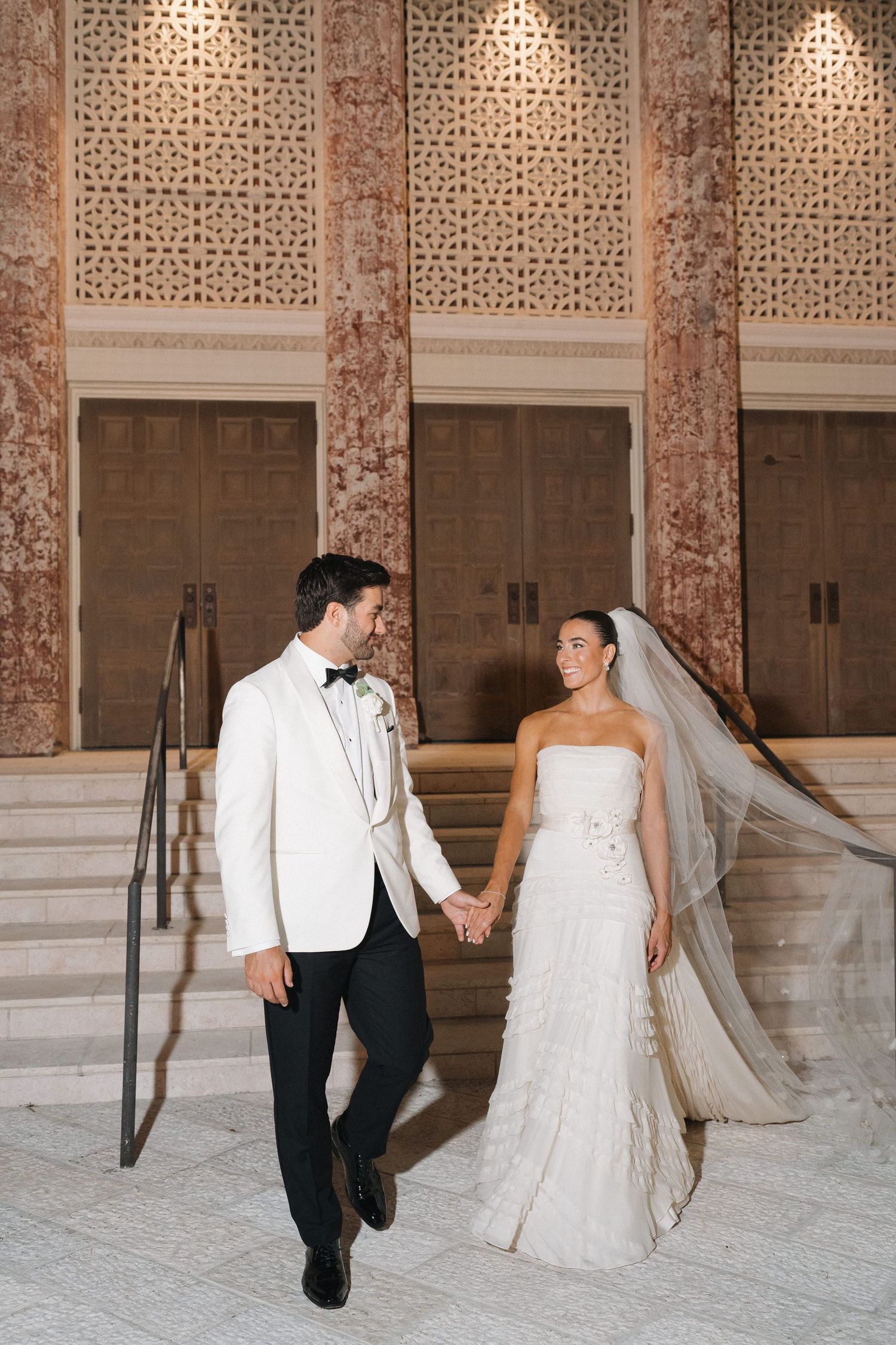 Newlyweds in formal attire descend marble steps with decorative columns and lighting.