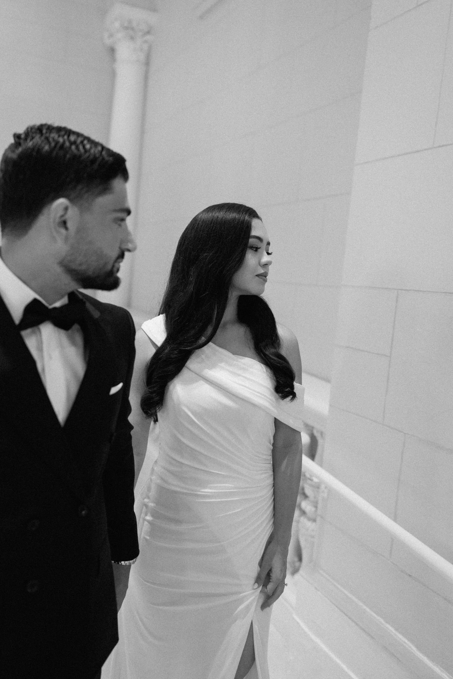 An elegant couple in formal attire poses against a white interior wall with classical columns.