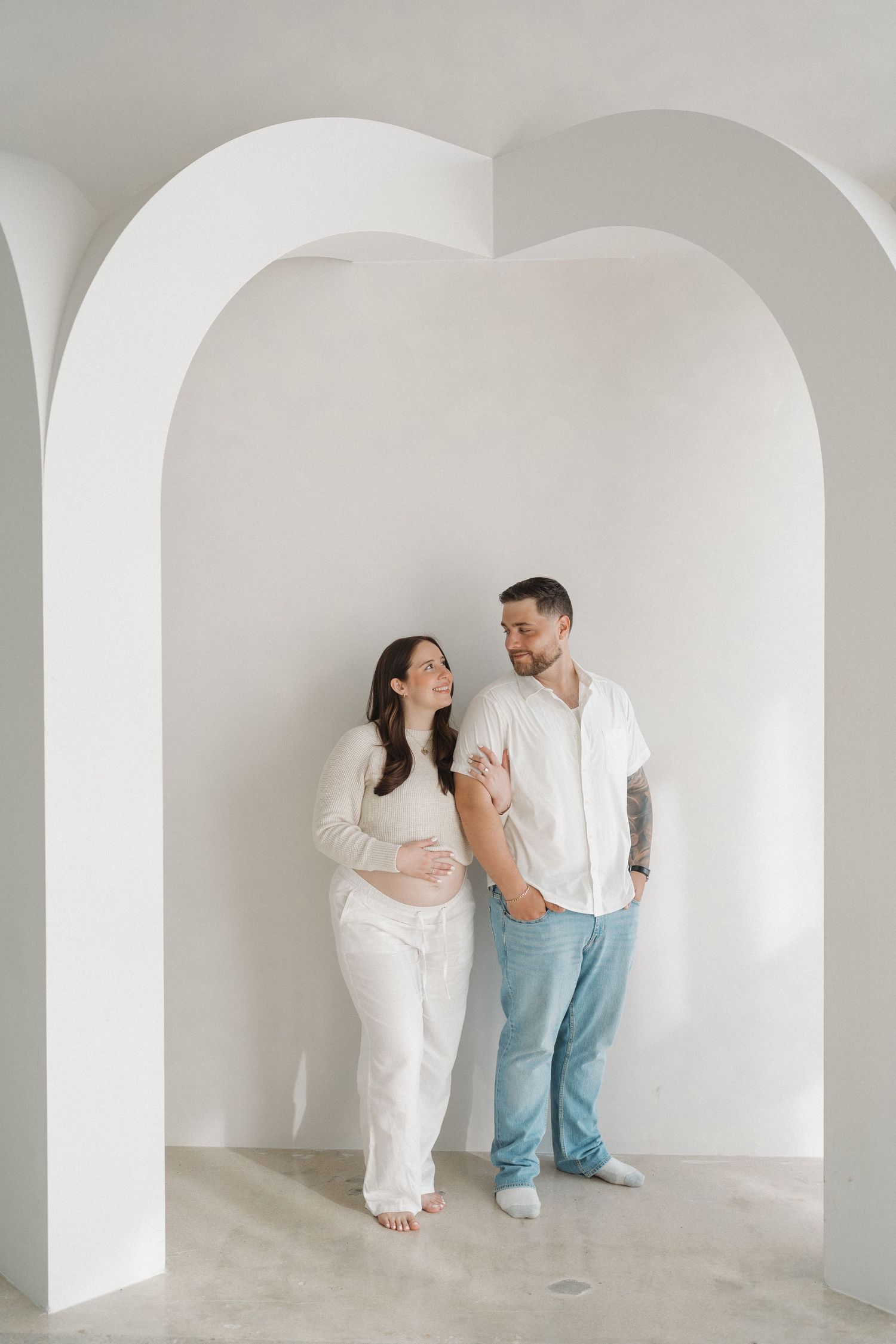 Couple sharing joyful moment under white archway during maternity photoshoot.