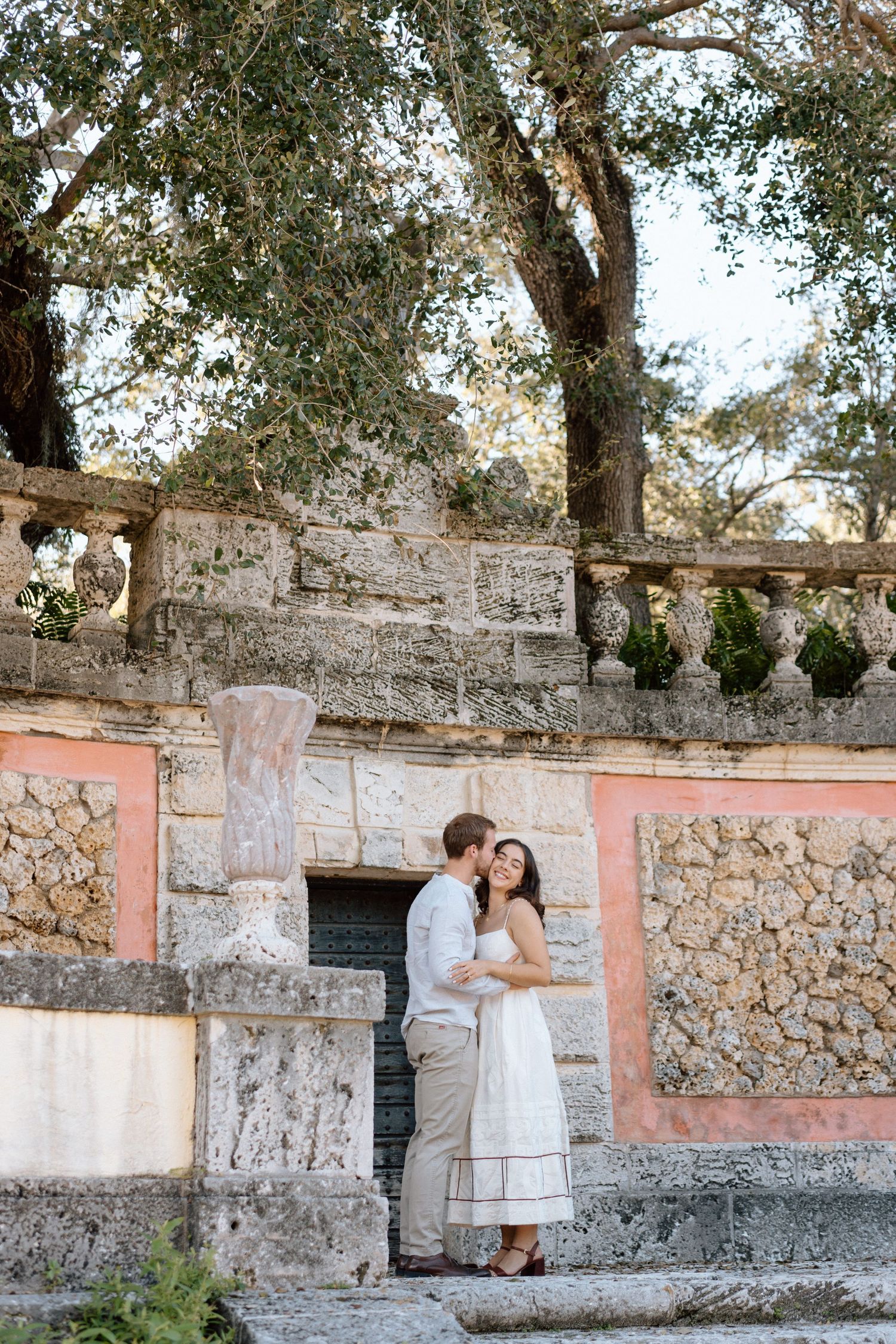 A romantic couple poses against a stone wall with pink accents under olive trees in a garden setting.