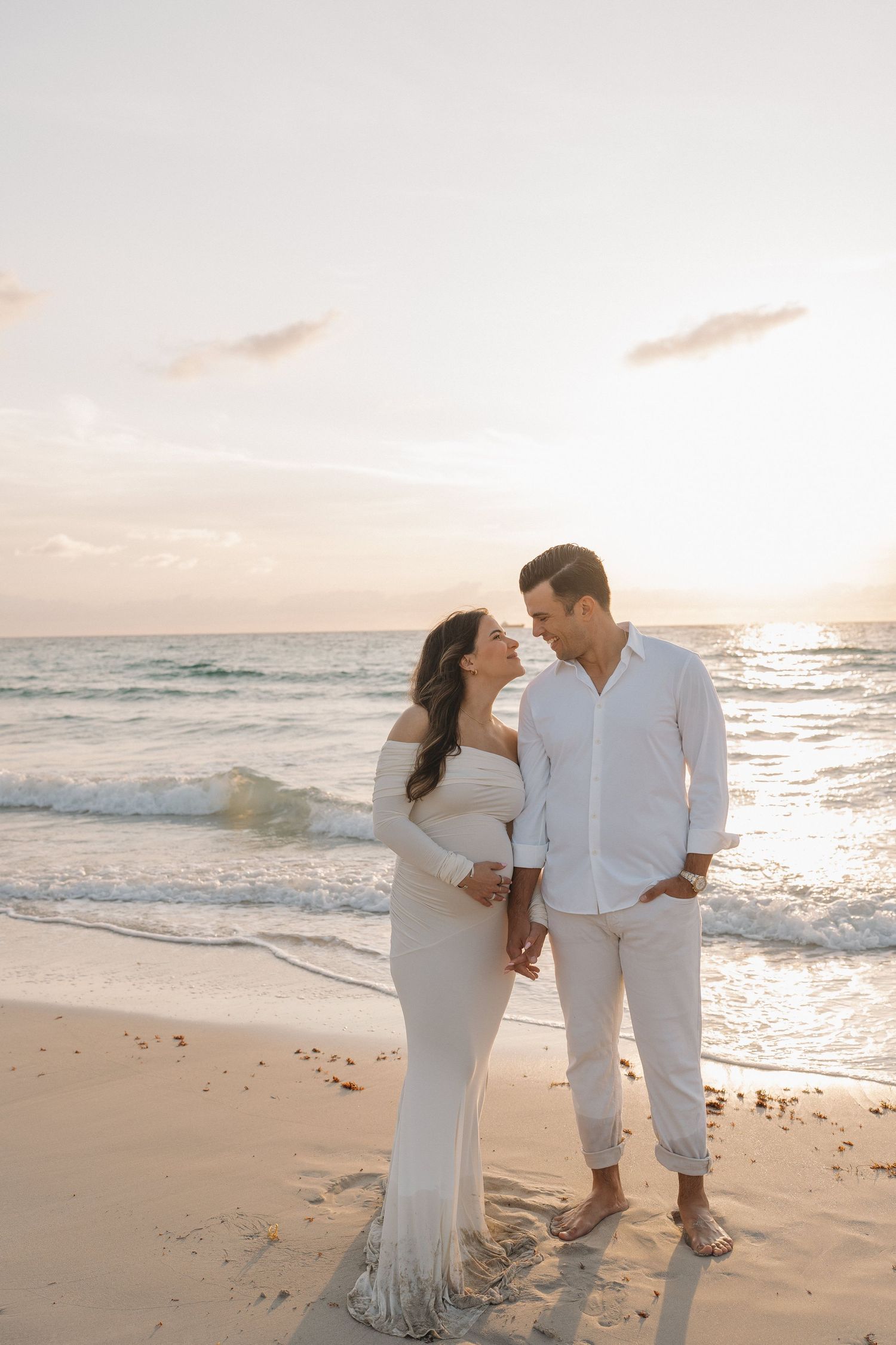 Couple in coordinated white outfits poses lovingly on a sandy beach during golden hour.