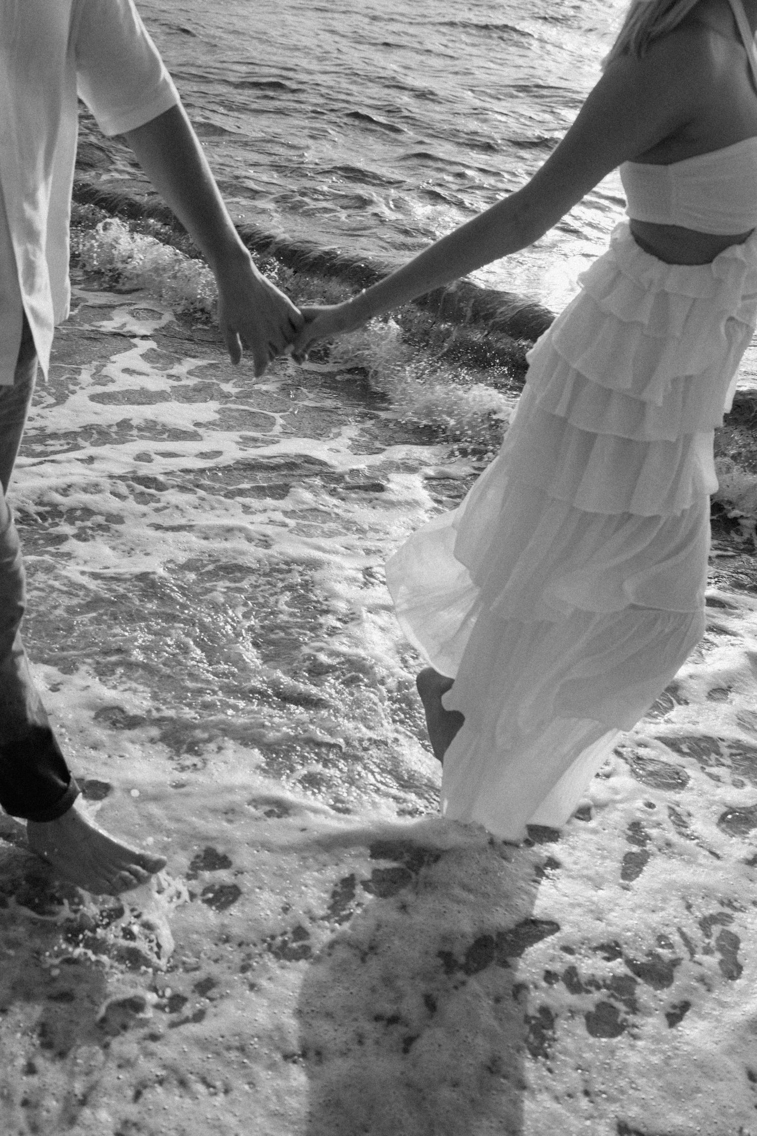 Black and white photo of hands held together as waves crash around feet on a beach.