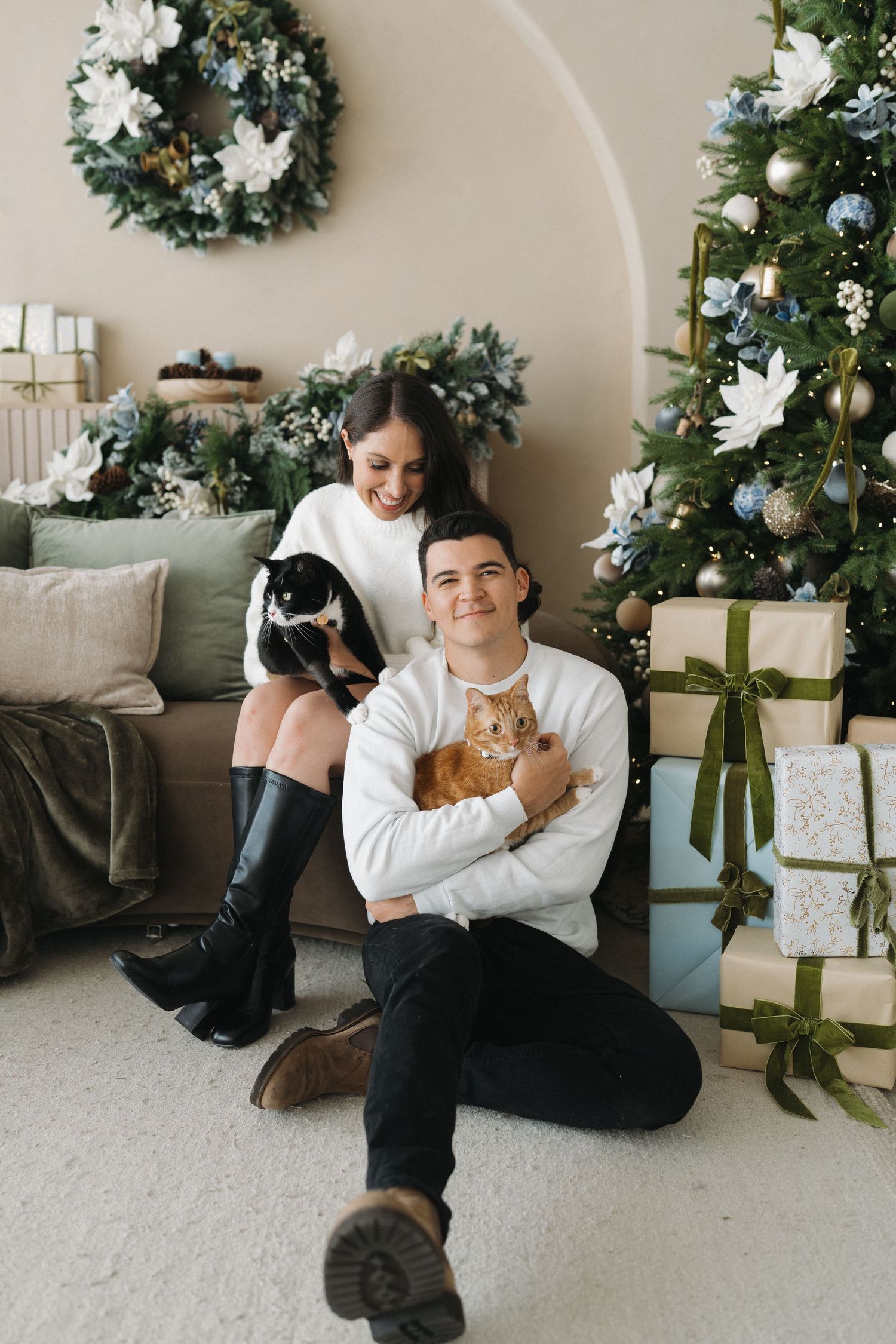 A couple poses with cats near a decorated Christmas tree and wrapped gifts in a cozy holiday living room.
