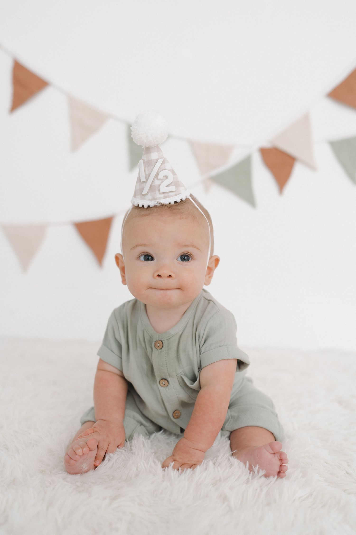 Baby wearing party hat sits on fluffy rug with festive bunting backdrop.