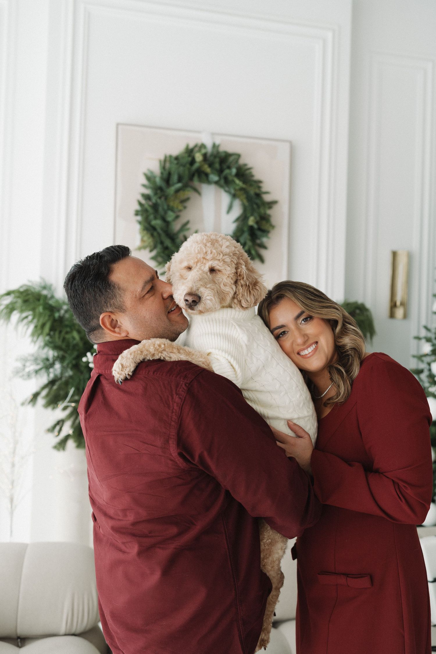 A couple in matching burgundy outfits holds a fluffy goldendoodle in a white sweater near a Christmas wreath.