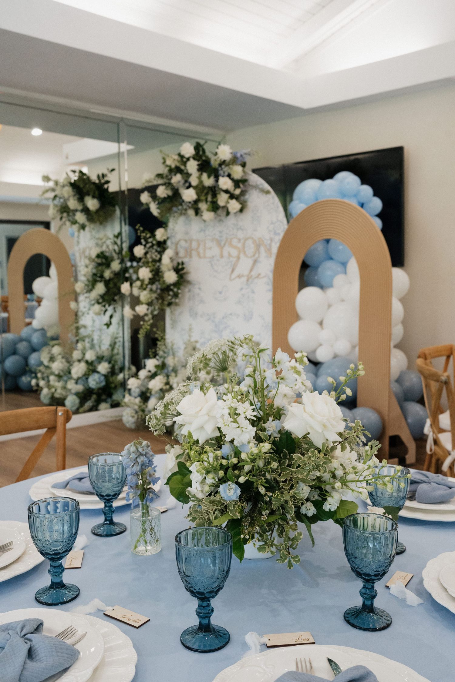 Elegant table setting with blue goblets, white floral centerpieces and tan arched backdrop for special event.