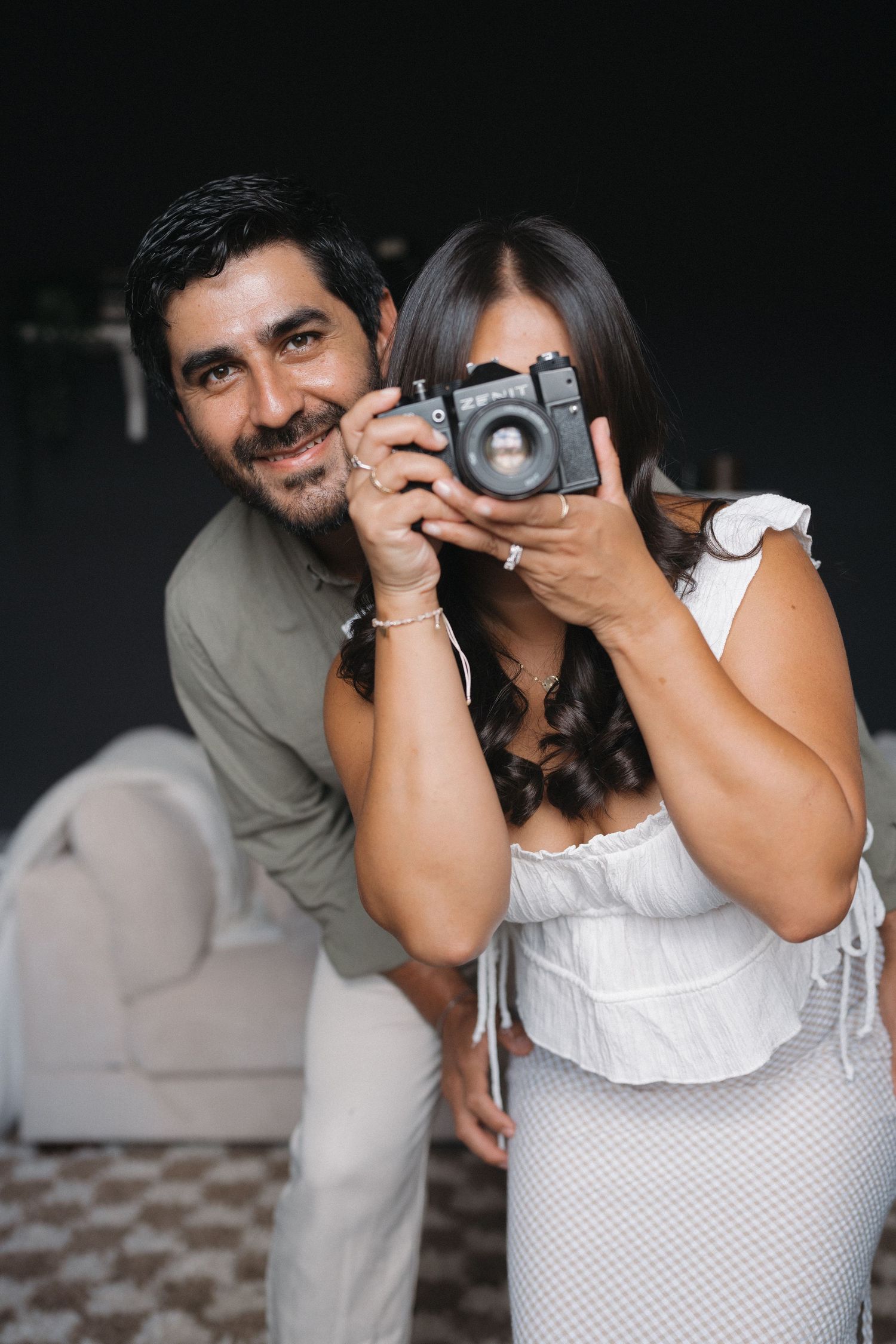 Playful moment of couple with vintage camera against dark wall background.