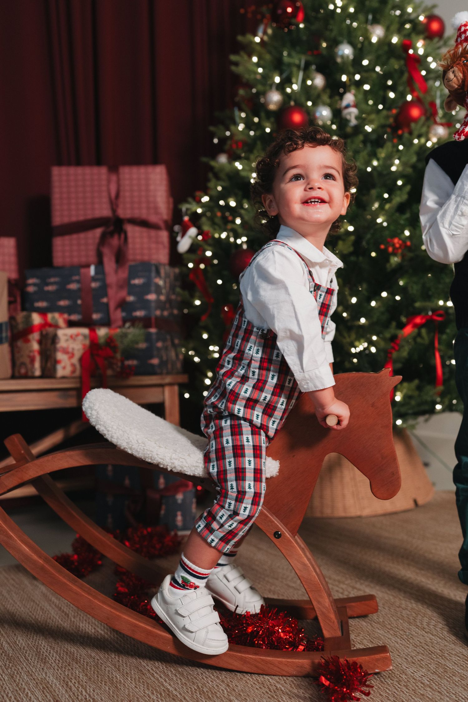 Happy toddler boy riding a wooden rocking horse in front of a decorated Christmas tree with gifts.
