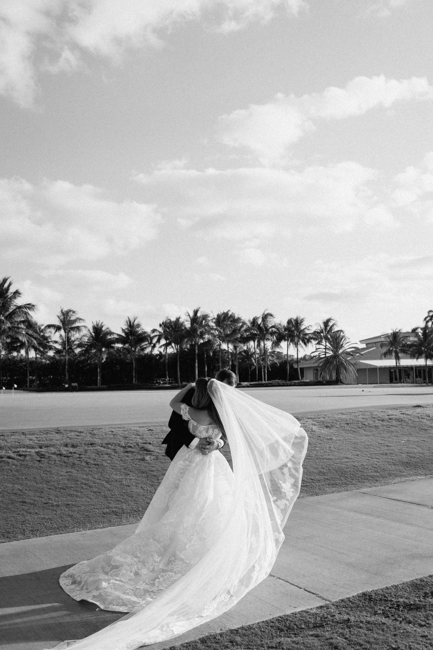 Black and white wedding photo with dramatic flowing veil against palm trees and sky.