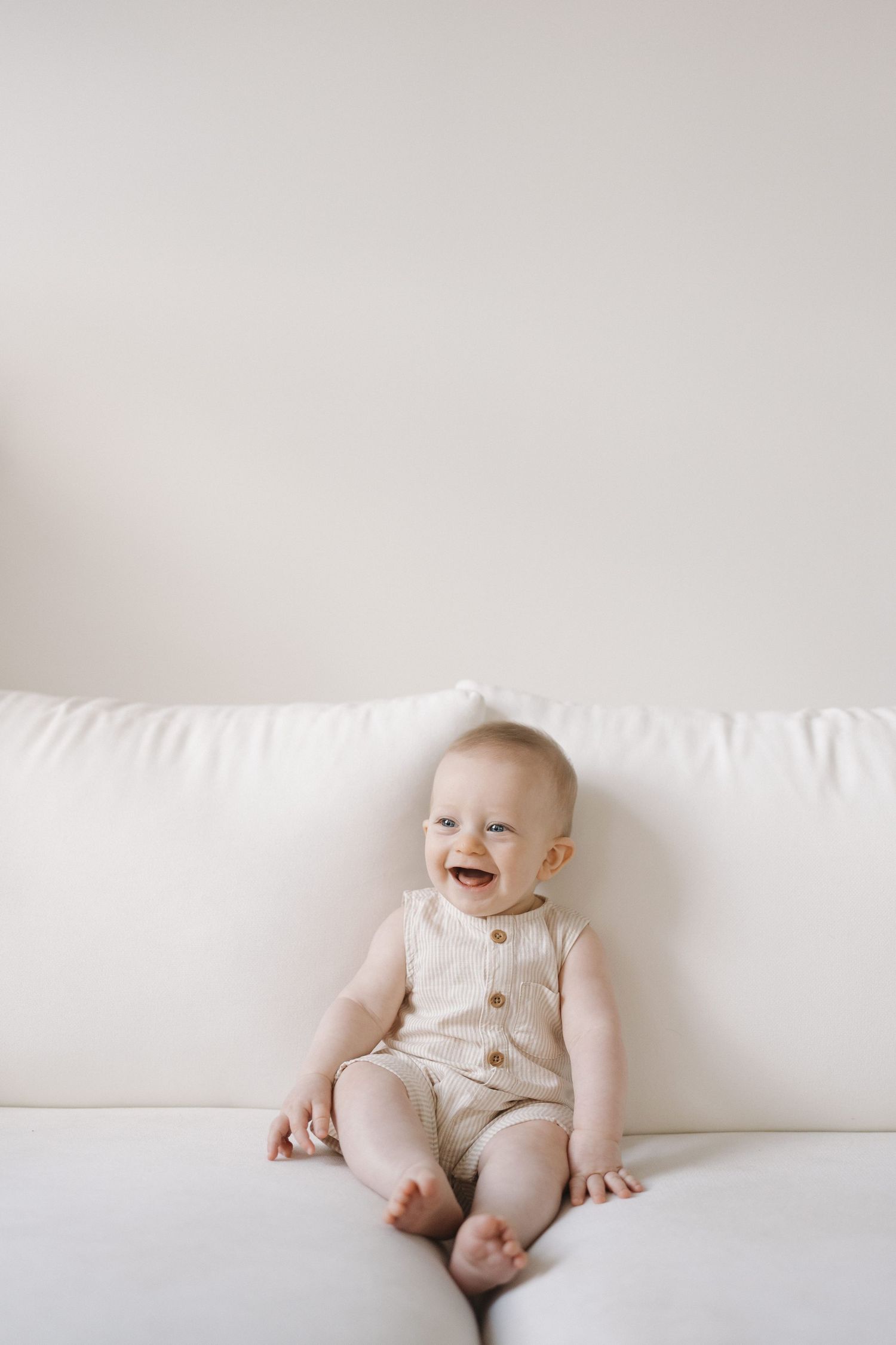 Happy baby in beige romper sitting on white couch.