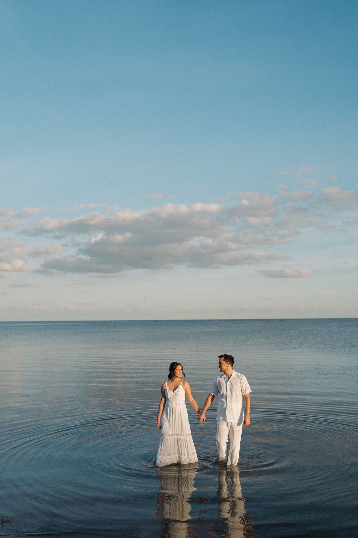 Couple stands together in calm waters during sunset with pastel clouds overhead.