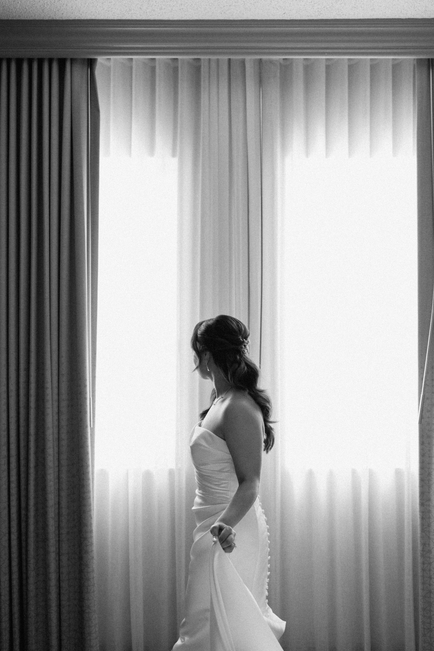 Black and white photo of bride silhouetted against curtained window.