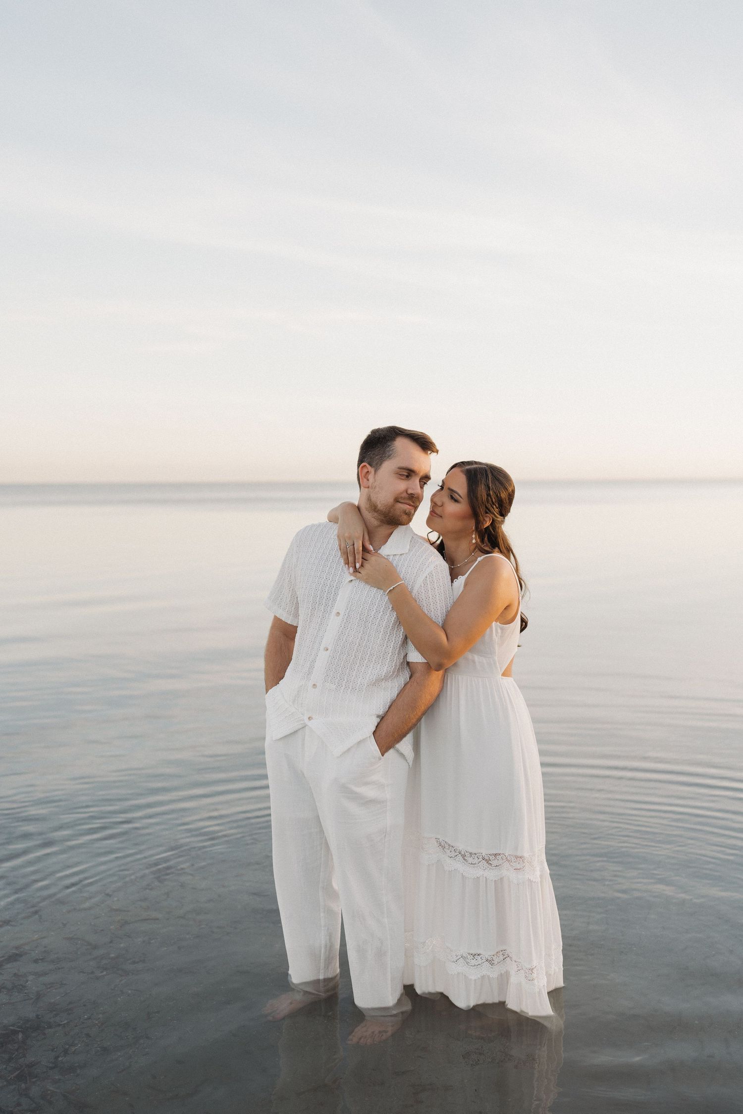 A couple in white clothing shares an intimate moment while standing in shallow ocean water.