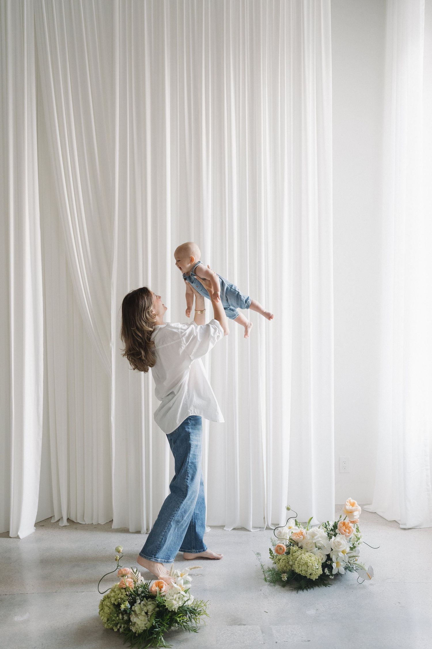 A joyful lifting moment against white curtains with floral arrangements on the floor.