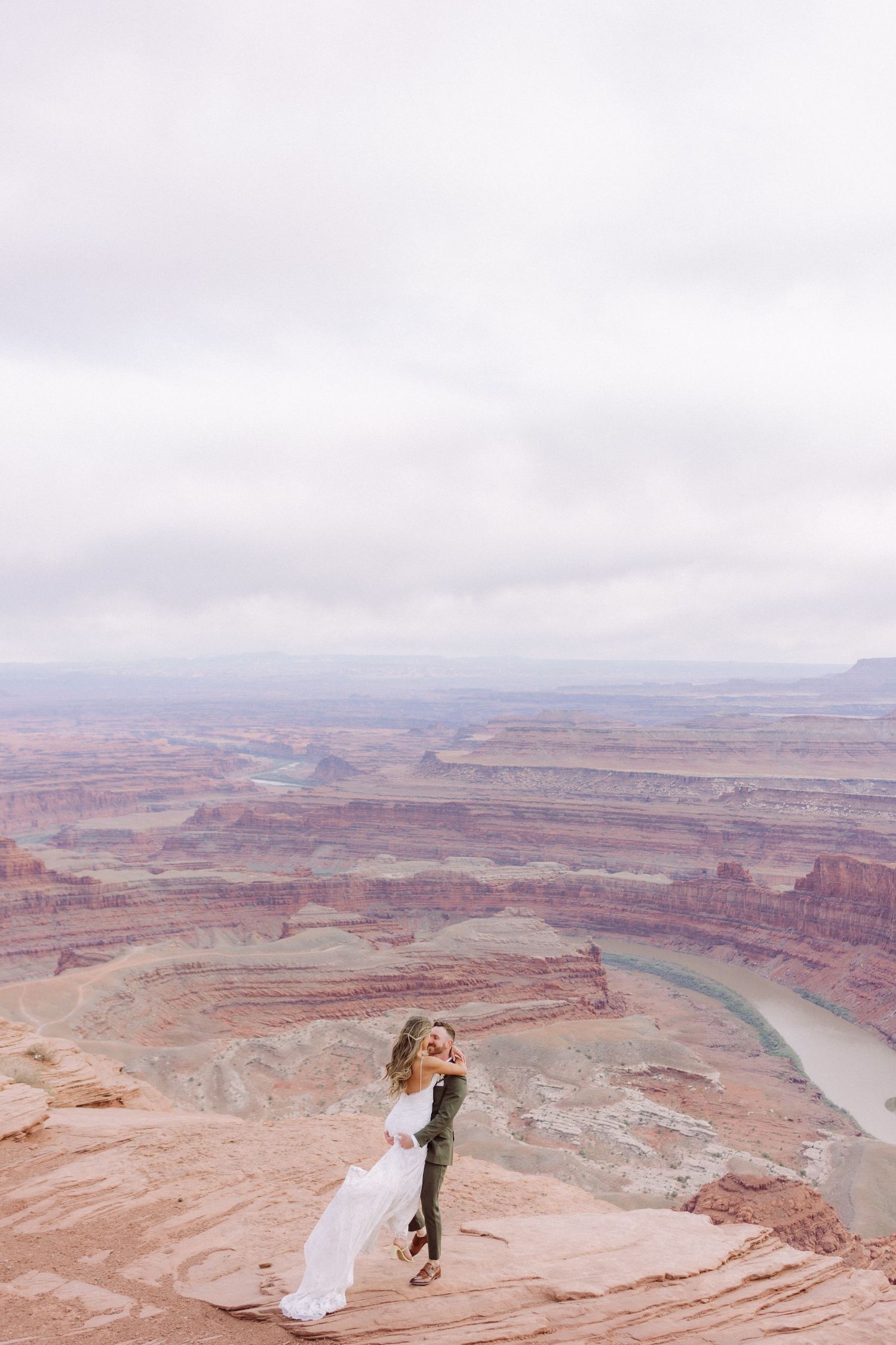 Bride in flowing dress poses dramatically on cliff edge overlooking desert canyon landscape.