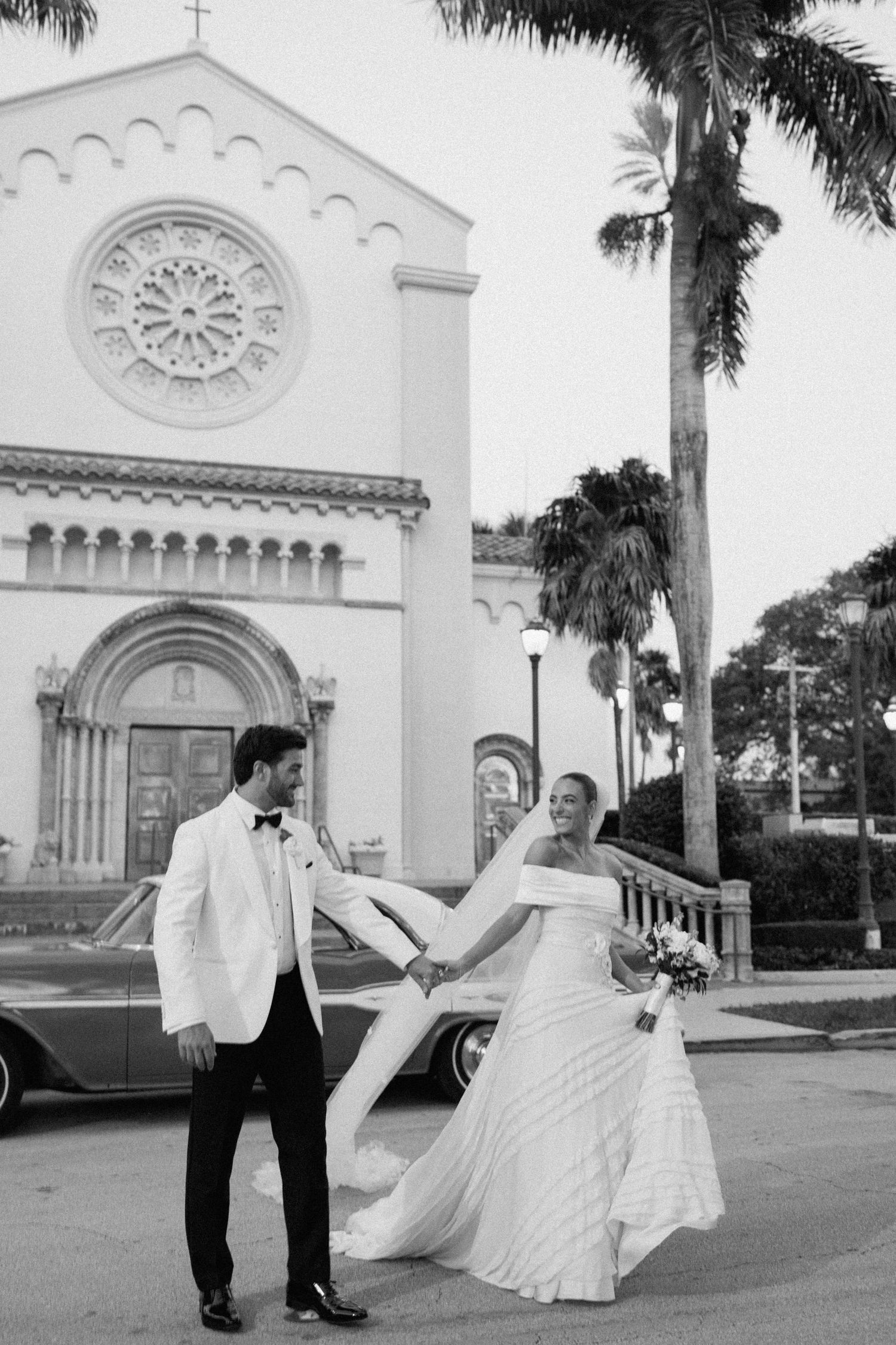 Black and white wedding portrait outside church with palm trees and vintage car.