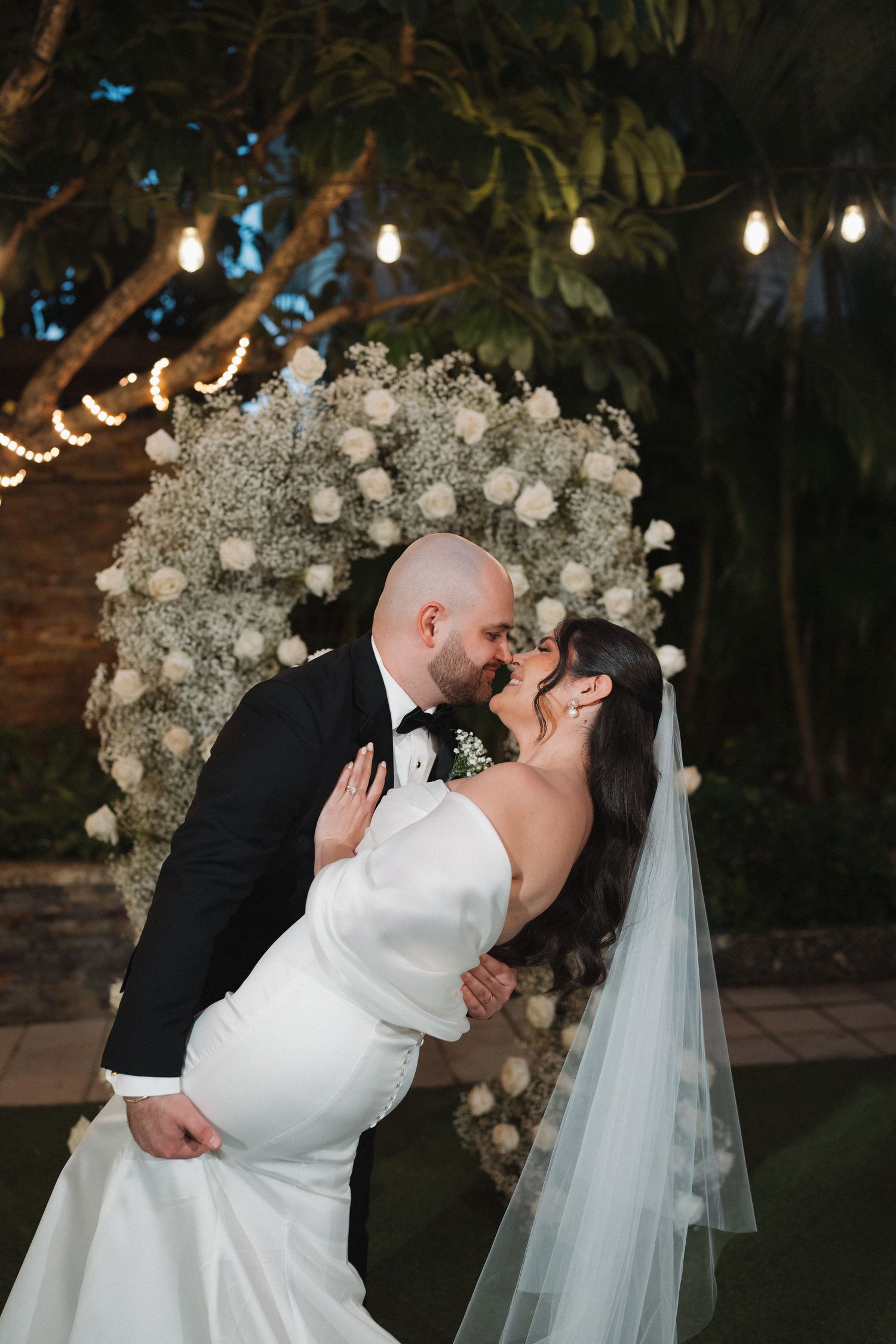 Wedding couple shares romantic moment under floral arch with string lights at night.