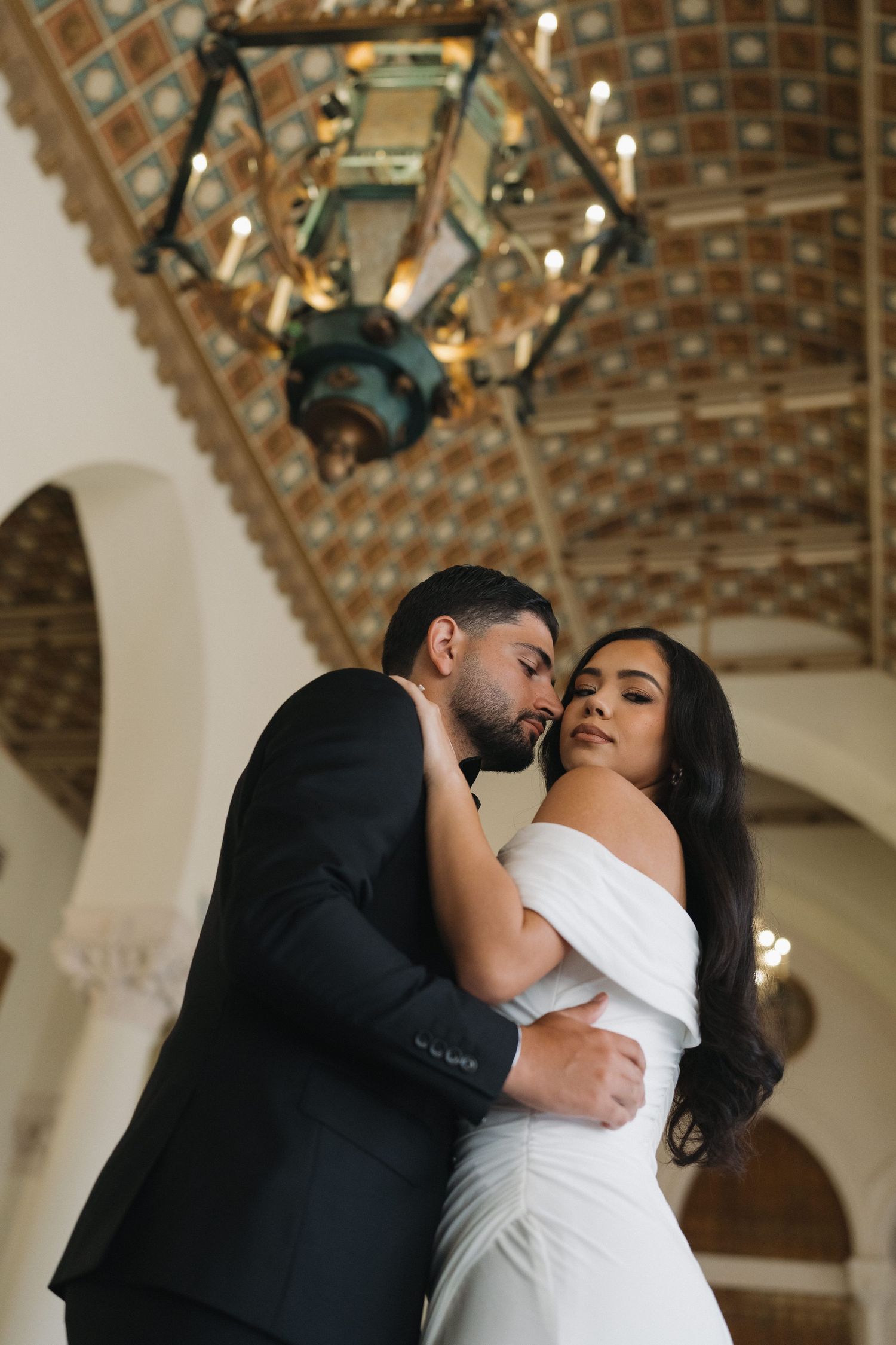 Intimate moment captured under an ornate chandelier in a classical architectural setting.