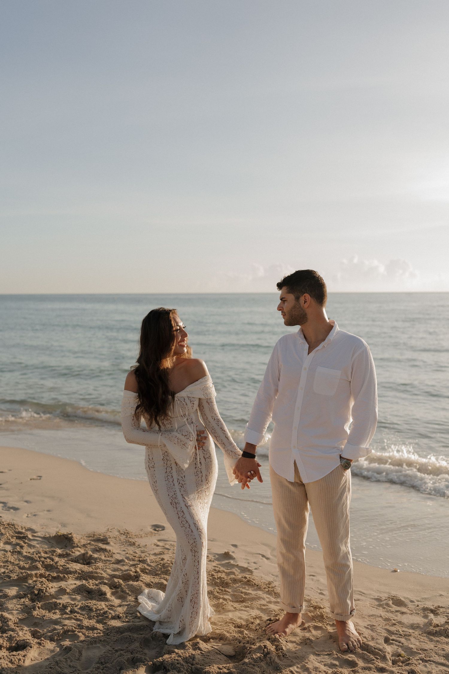 A couple in coordinated white outfits walks hand in hand along the sandy shoreline.