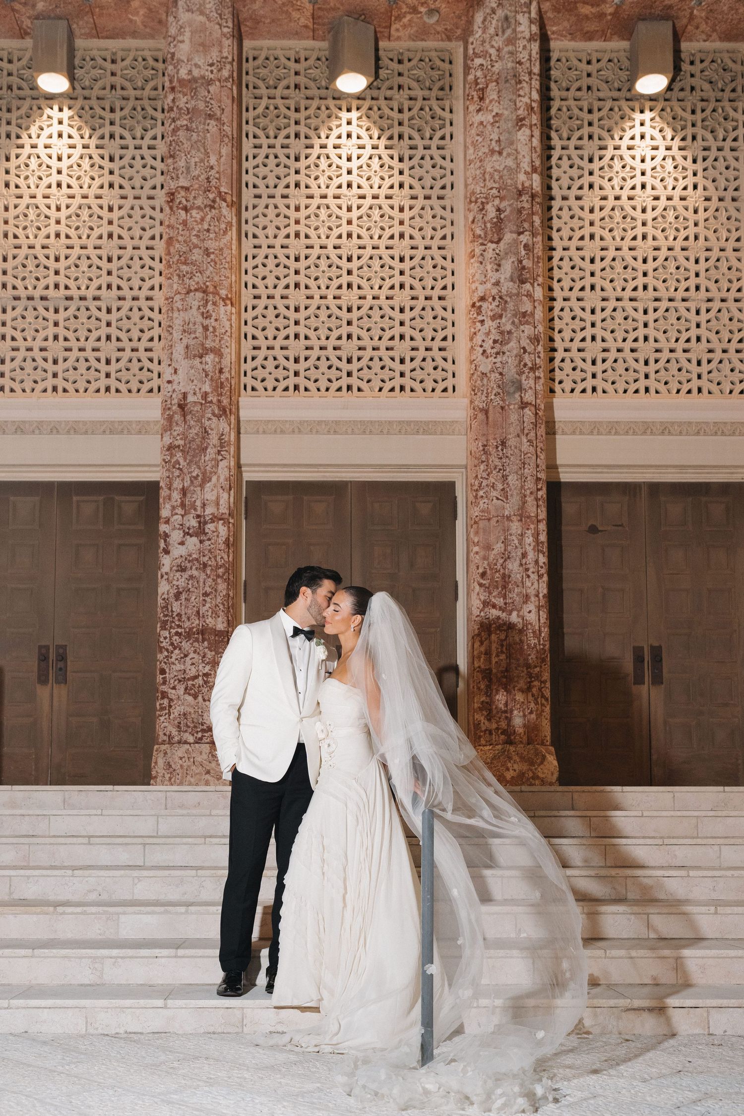 Elegant wedding couple embraces on marble steps beneath ornate architectural details and spotlights.