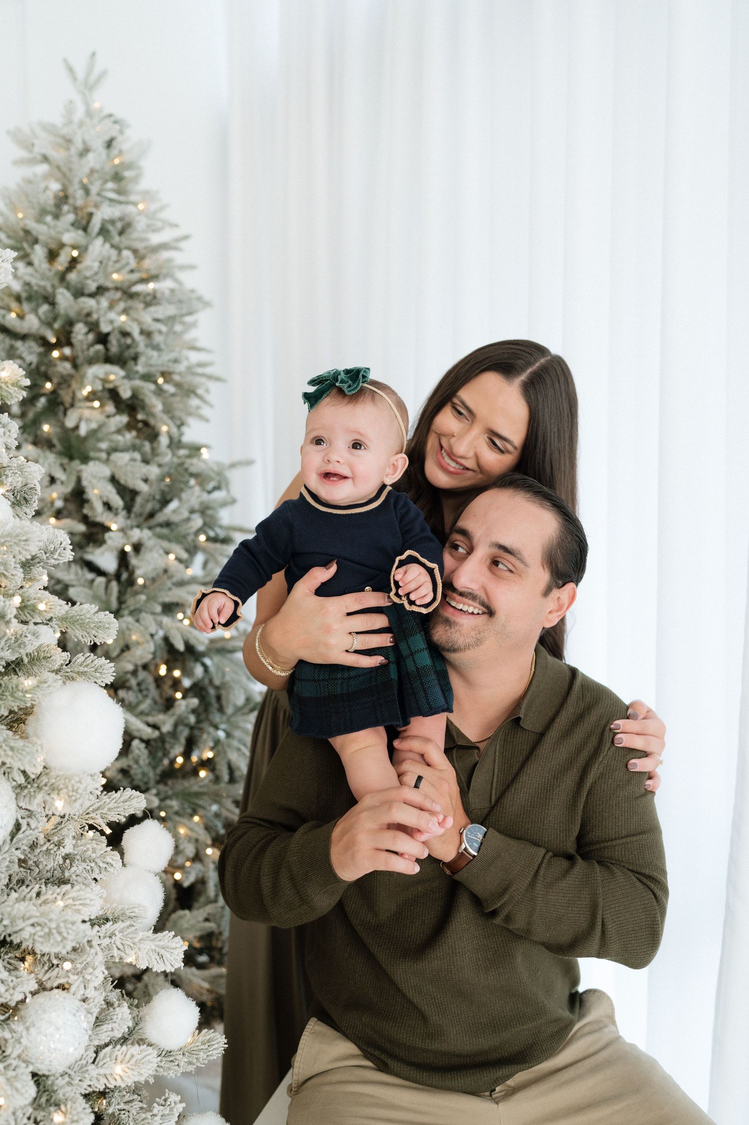 Happy family with baby girl in navy dress poses by decorated Christmas tree in white room.