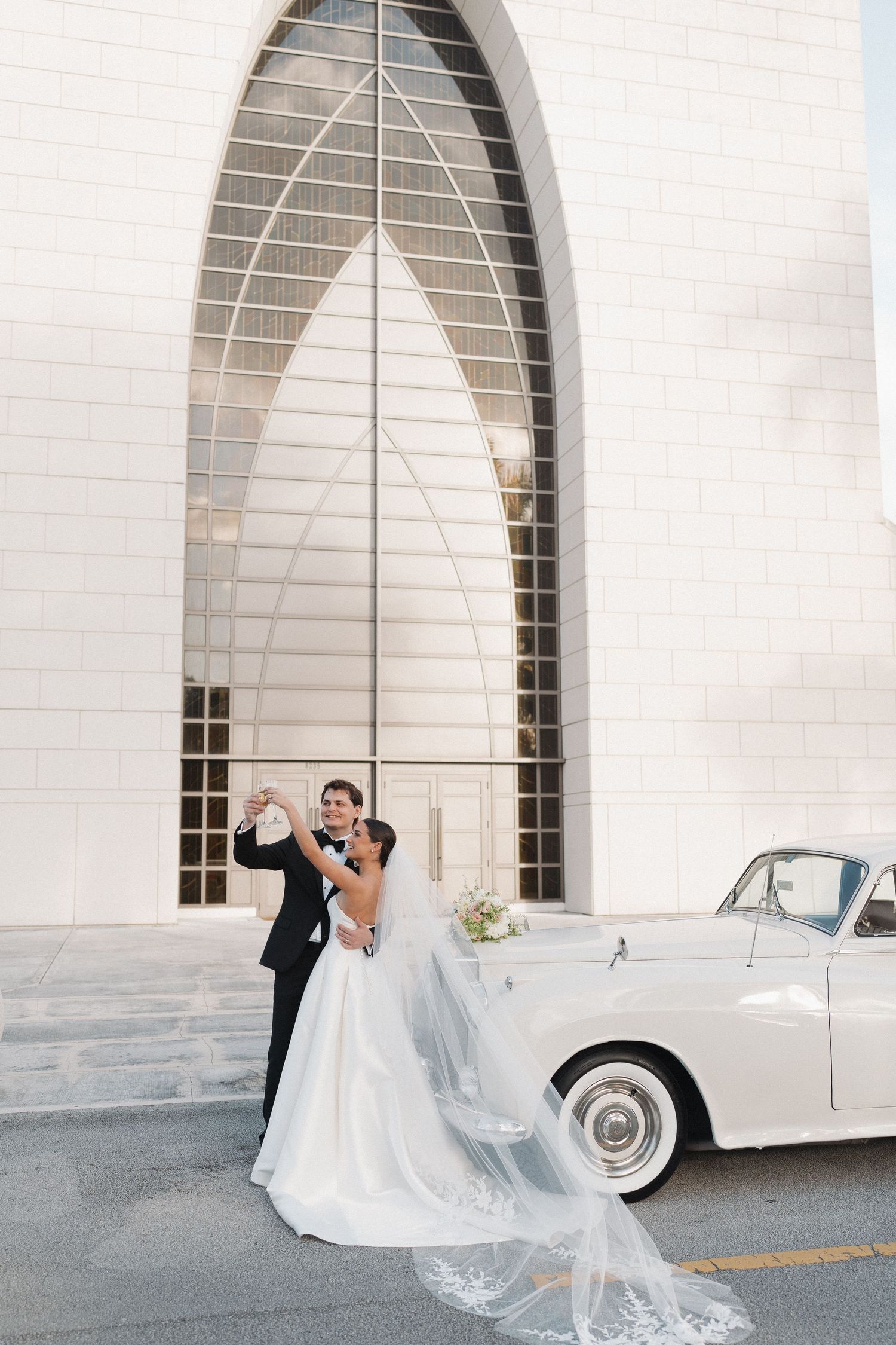 Newlyweds pose elegantly by a classic white car in front of a modern church archway.