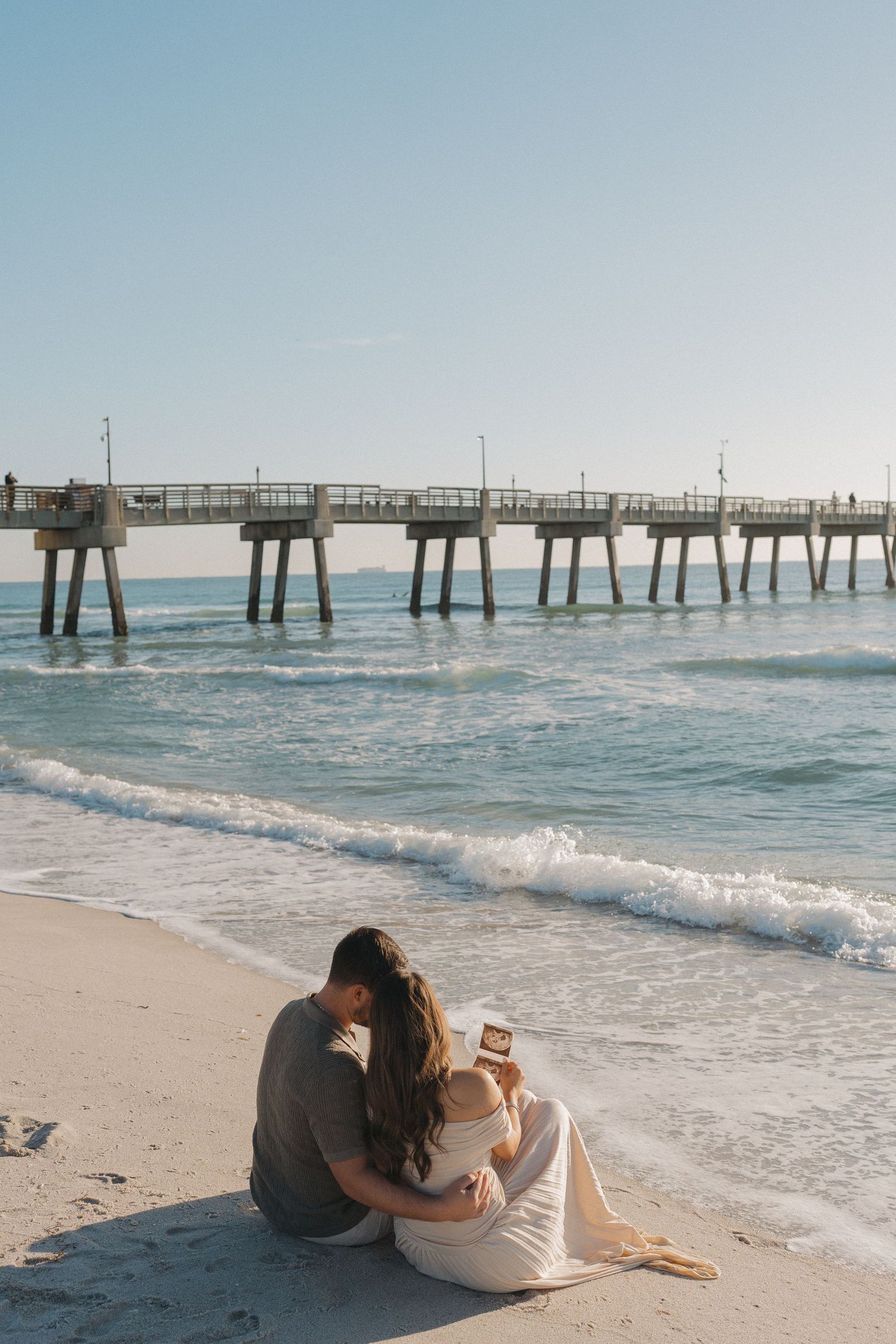A couple sits together on a beach watching waves roll in near a wooden pier during sunset.