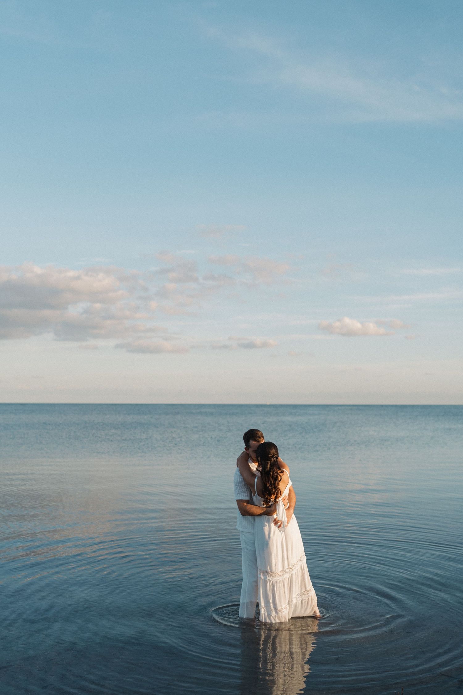 A couple embraces while standing in calm ocean waters at sunset with pastel clouds in the background.