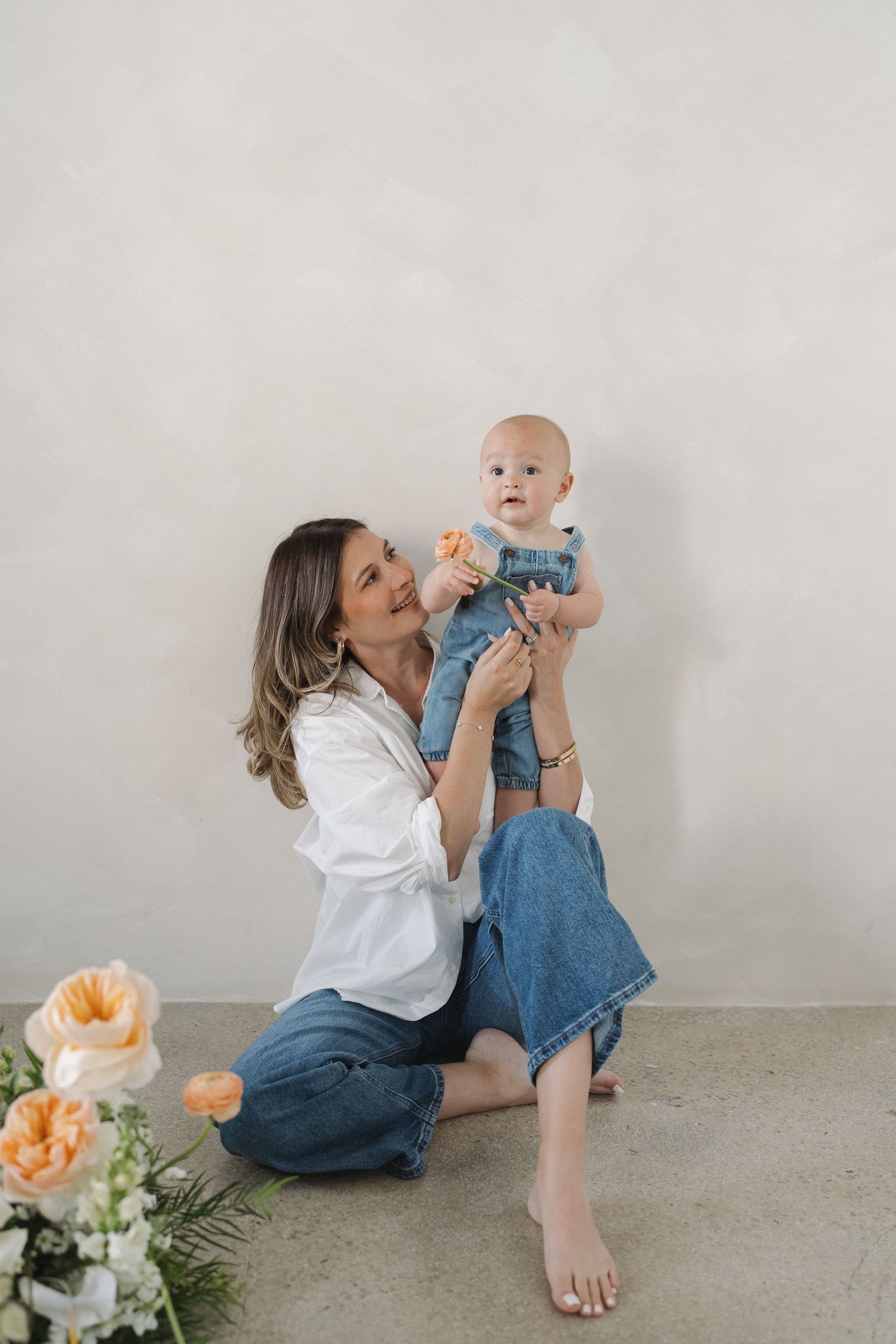 A tender moment captured with a baby in denim against a light grey background with peach-colored flowers.
