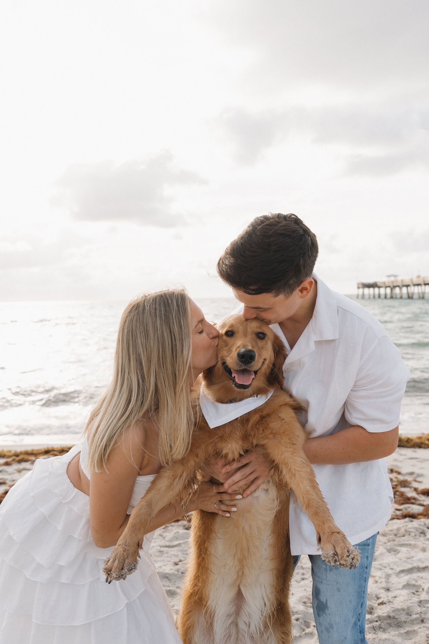 Couple kissing their happy golden retriever on a sunny beach with pier in background.