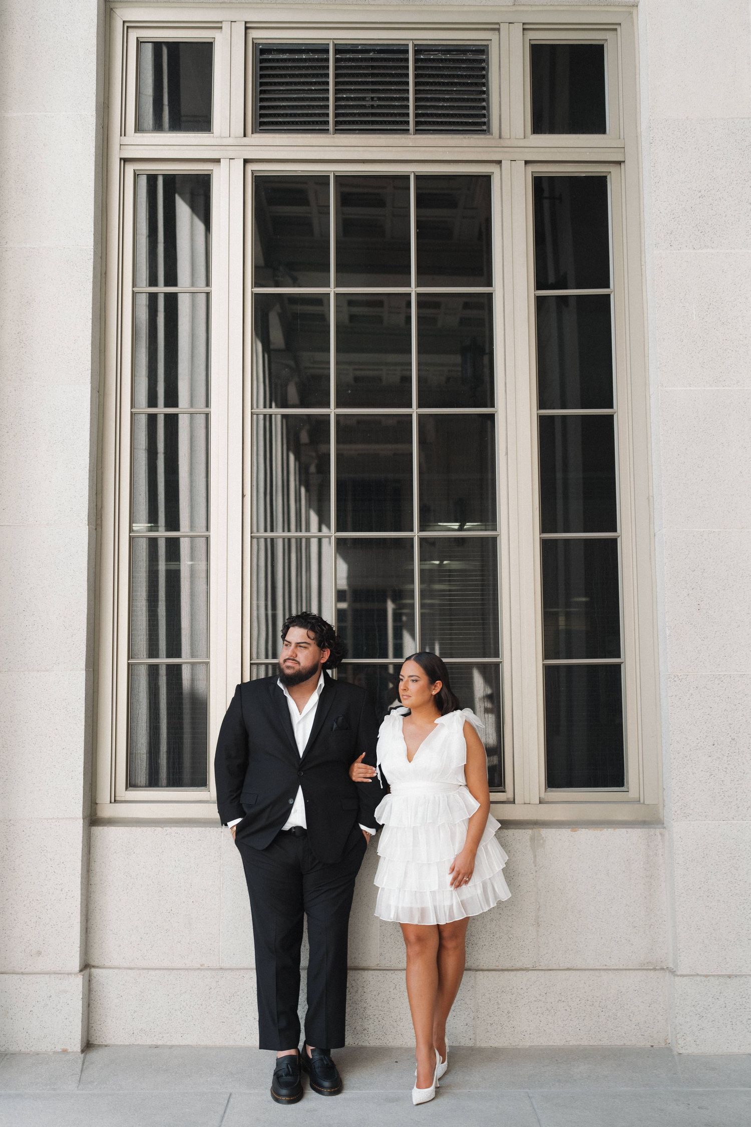 A couple in contrasting formal wear poses against a grand building window.