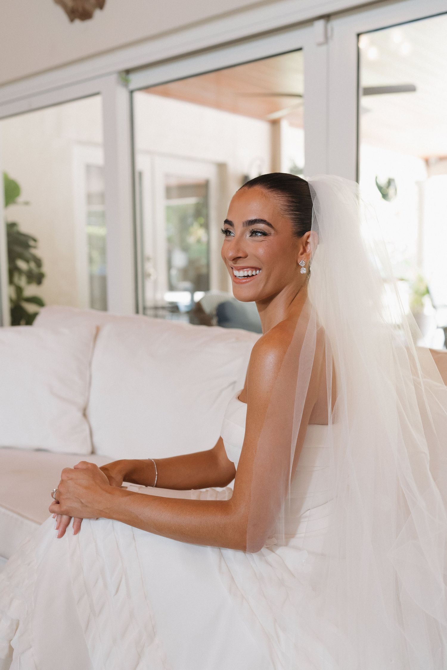 Bride wearing veil sits on white couch with joyful expression.
