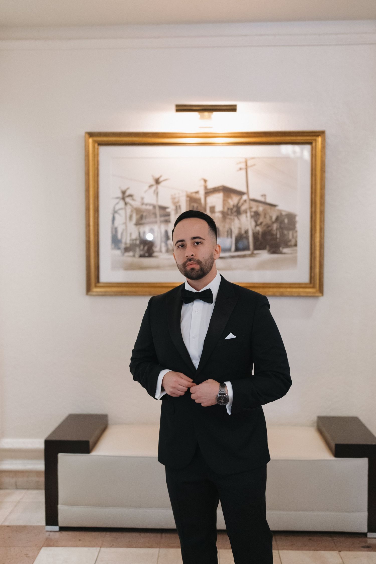 Wedding day portrait of groom in classic tuxedo standing in front of framed artwork.