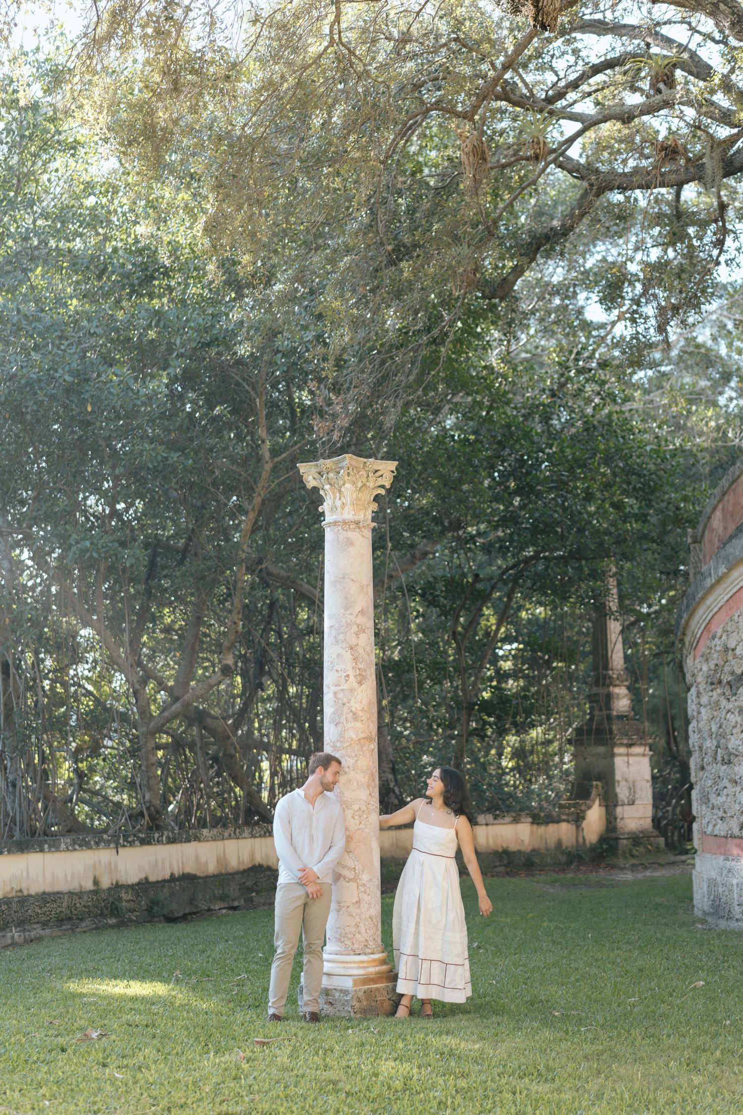 A couple stands beside a classical stone column in a sunlit garden setting.