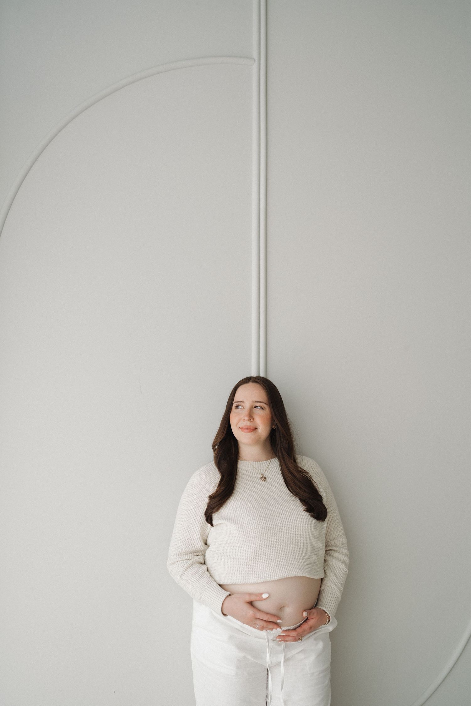 Minimalist maternity portrait of woman in cream sweater against geometric wall design.